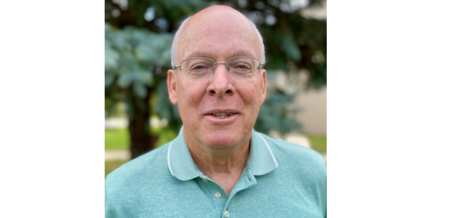 Portrait of a middle-aged man with glasses, wearing a light blue polo shirt, outdoors with trees in the background.