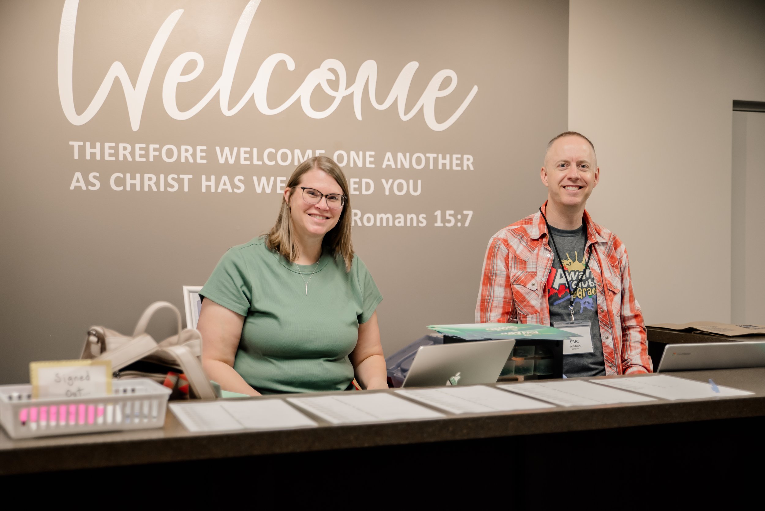 Two smiling people, a woman with glasses and a man, sitting behind a table at a welcome booth with a large welcome sign on the wall behind them and a Bible verse from Romans 15:7.