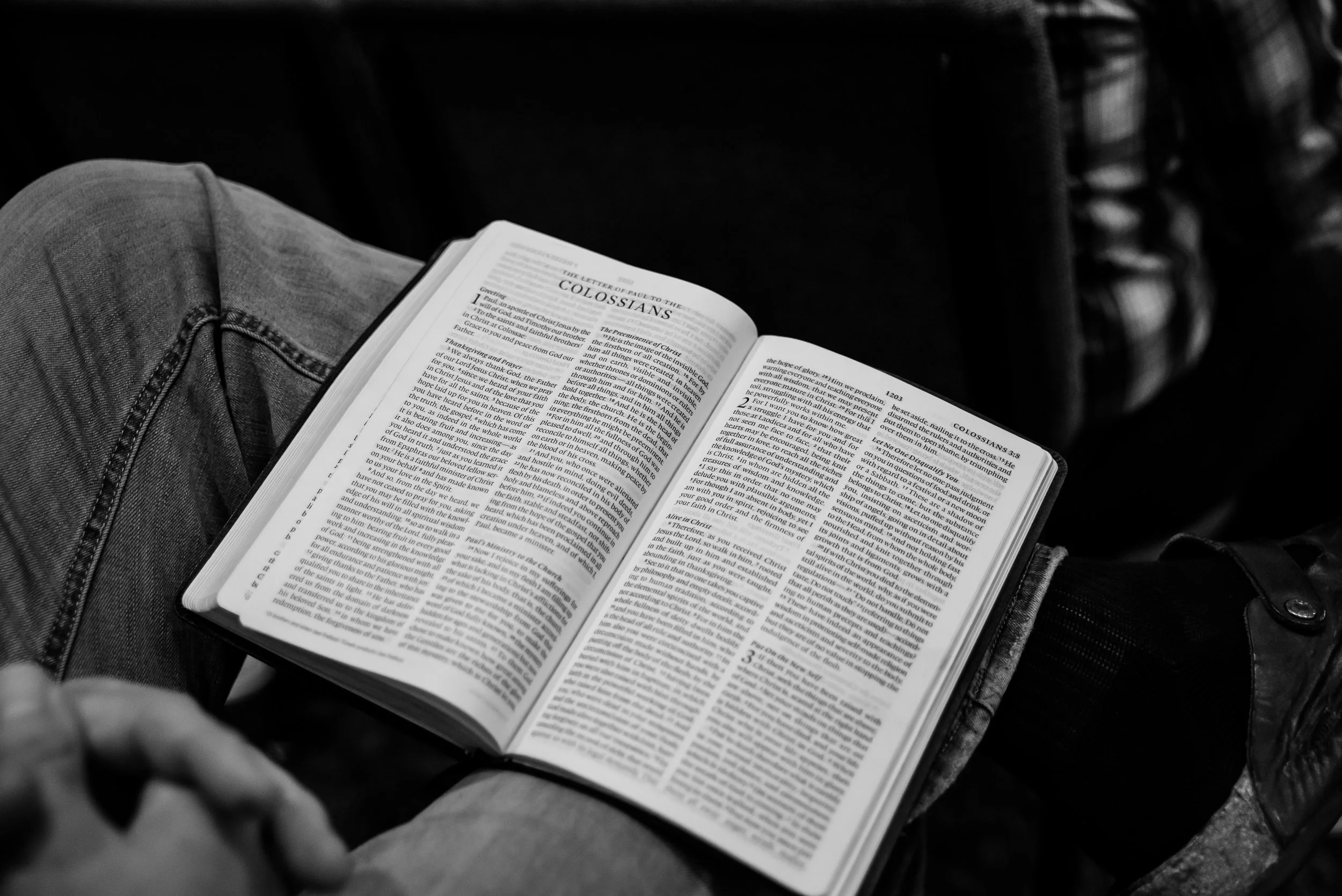 Person wearing jeans sitting and reading the Bible, specifically the book of Colossians, in a dark setting.