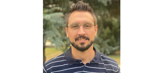 A man with glasses, a beard, and short beard in a navy and white striped polo shirt, standing outdoors with trees in the background.