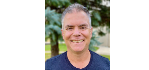 A smiling man with gray hair wearing a dark blue shirt outdoors with trees in the background.