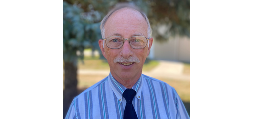 A middle-aged man with glasses, wearing a striped blue shirt and a dark tie, smiling outdoors with trees and a building in the background.