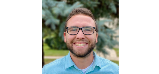 A man with glasses and a beard smiling outdoors, wearing a blue button-up shirt, with trees in the background.