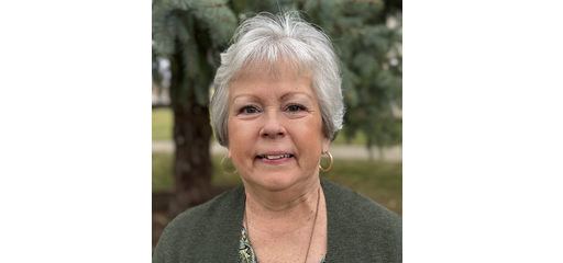 An elderly woman with short gray hair smiling outdoors with trees in the background.