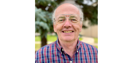 A smiling elderly man with glasses, wearing a red and blue plaid shirt, outdoors with greenery in the background.