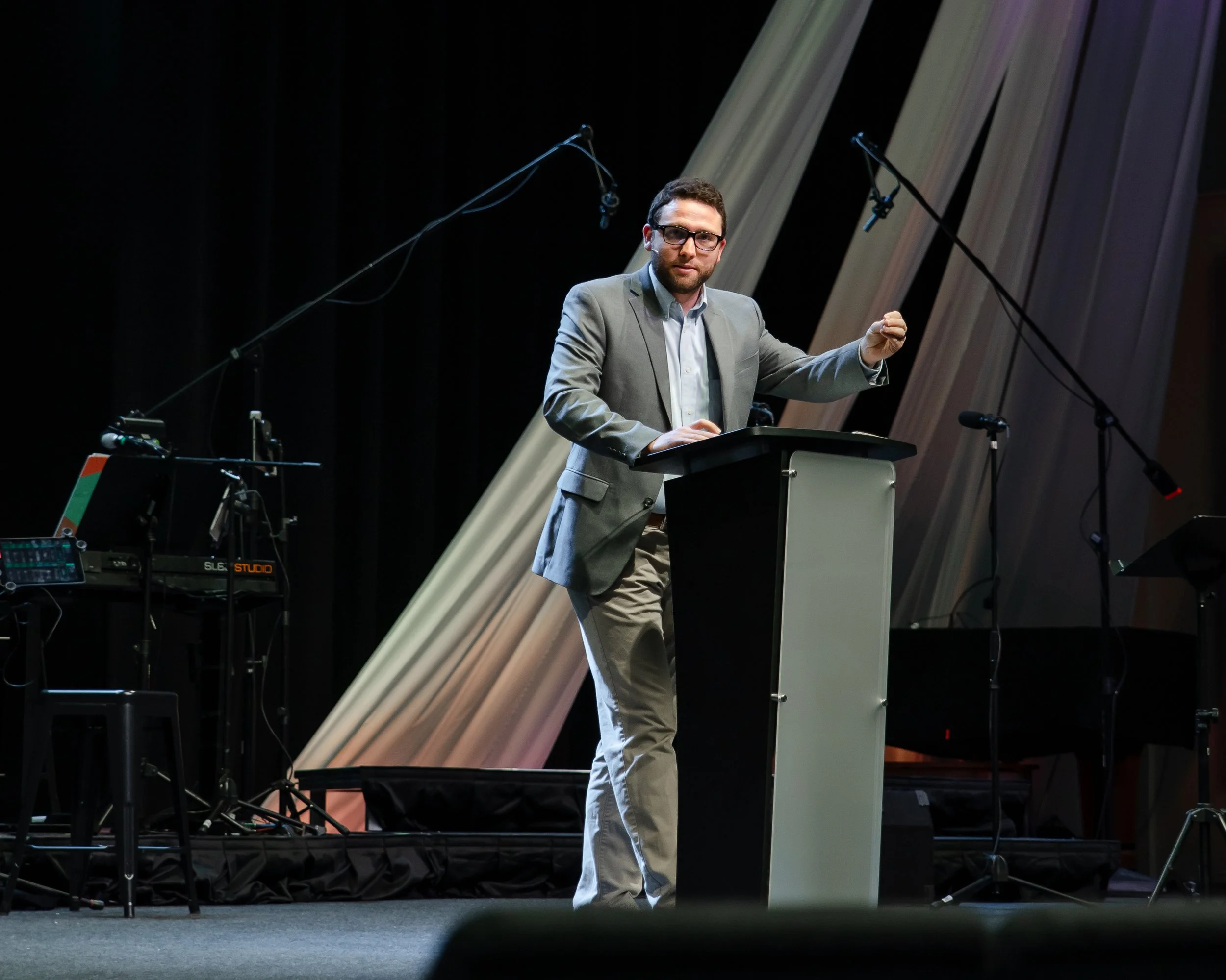 A man with glasses and a beard in a gray suit giving a presentation at a podium on a stage, with microphones and audio equipment around him.