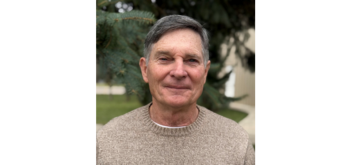 A middle-aged man with gray hair smiling outdoors, wearing a beige sweater, with trees in the background.