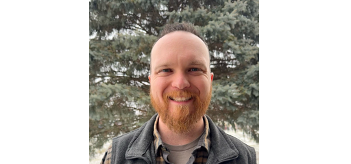 A smiling man with a beard and short hair outdoors in front of trees.