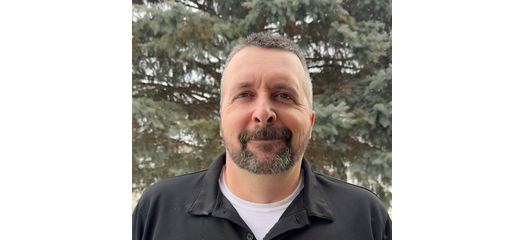 A man with short dark hair and a beard, wearing a black jacket over a white shirt, standing outdoors in front of trees.