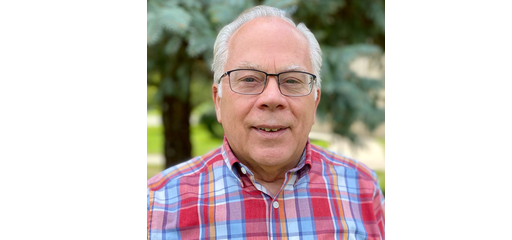 An older man with white hair, glasses, and wearing a plaid shirt, smiling outdoors with greenery in the background.