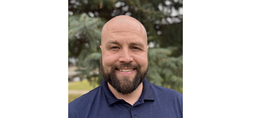 A smiling man with a beard and bald head wearing a blue collared shirt outdoors with trees in the background.