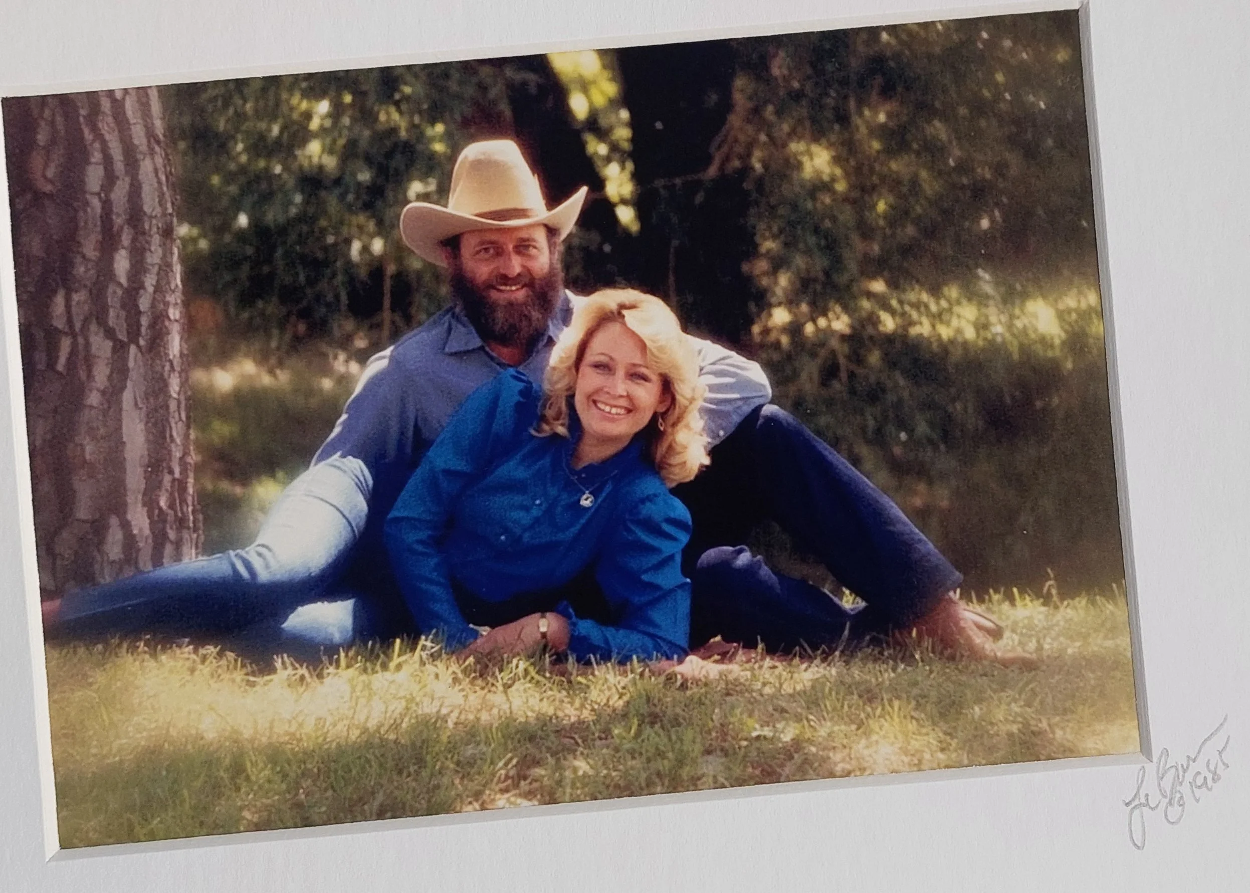A smiling man with a beard wearing a cowboy hat and a woman with blonde hair sitting on the grass in a park with trees in the background.