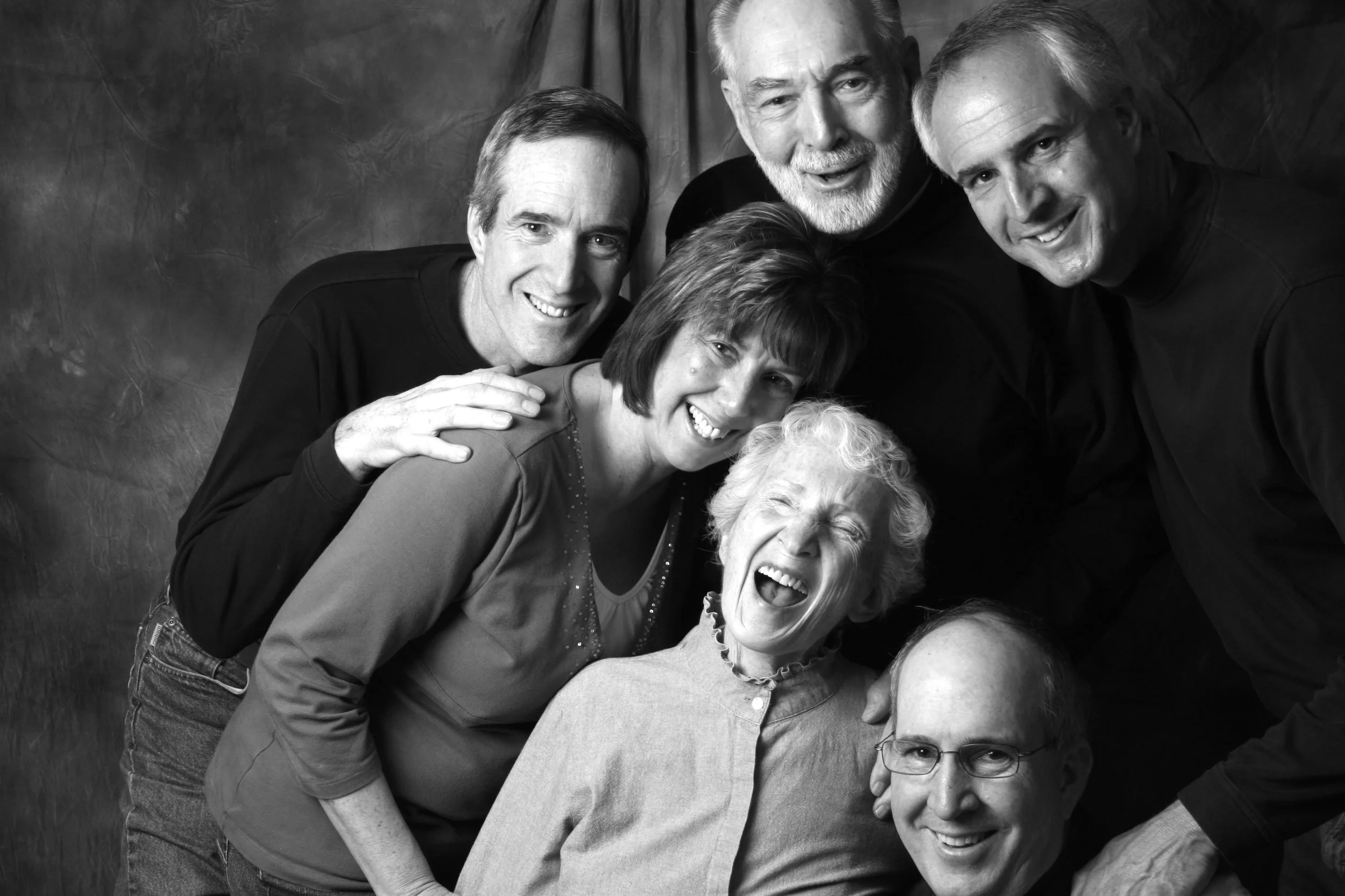A black-and-white photo of a group of six happy people, including elderly and middle-aged, smiling and posing closely together.
