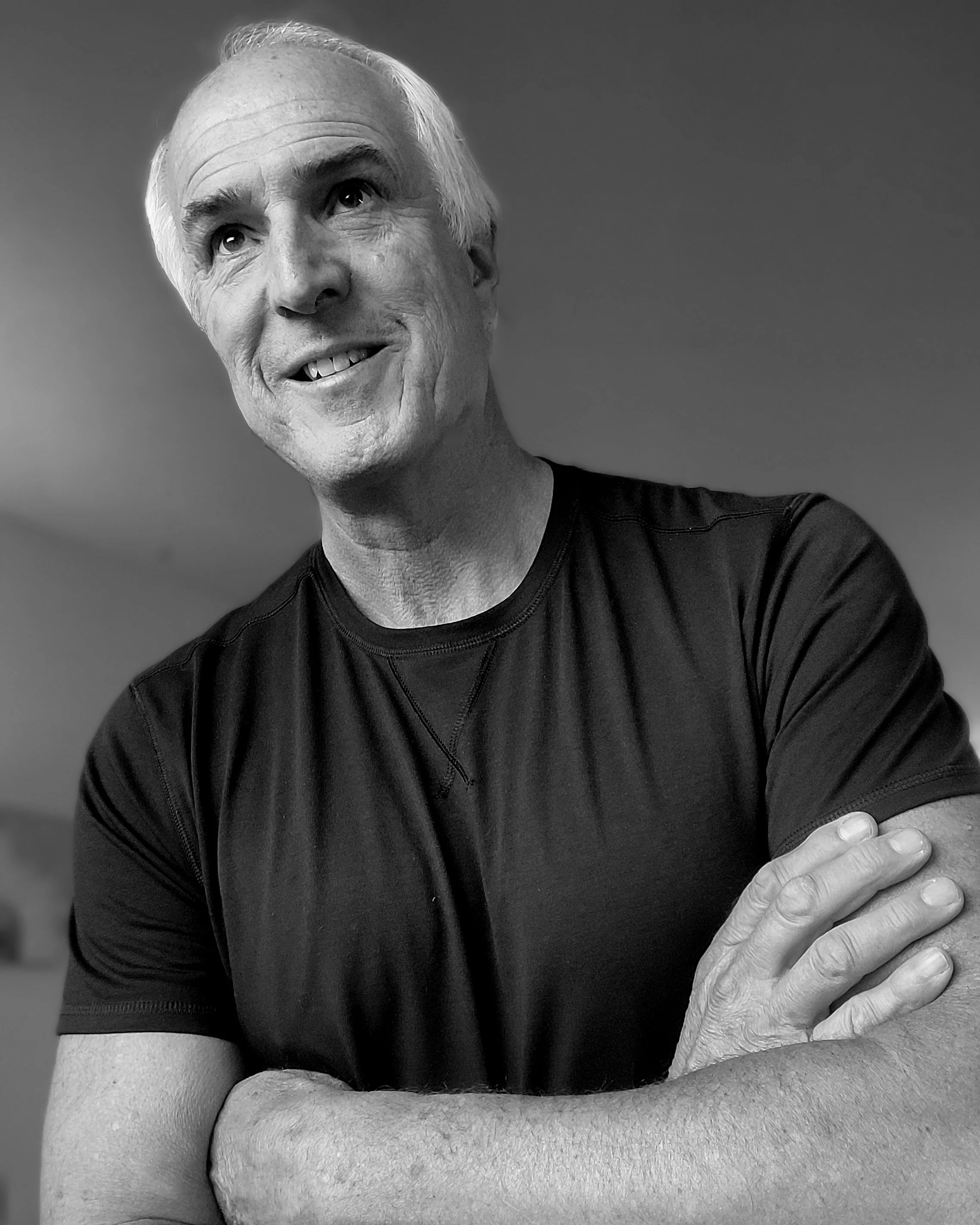 A black and white close-up of an older man with gray hair smiling, wearing a dark T-shirt, and resting his hand on his arm.