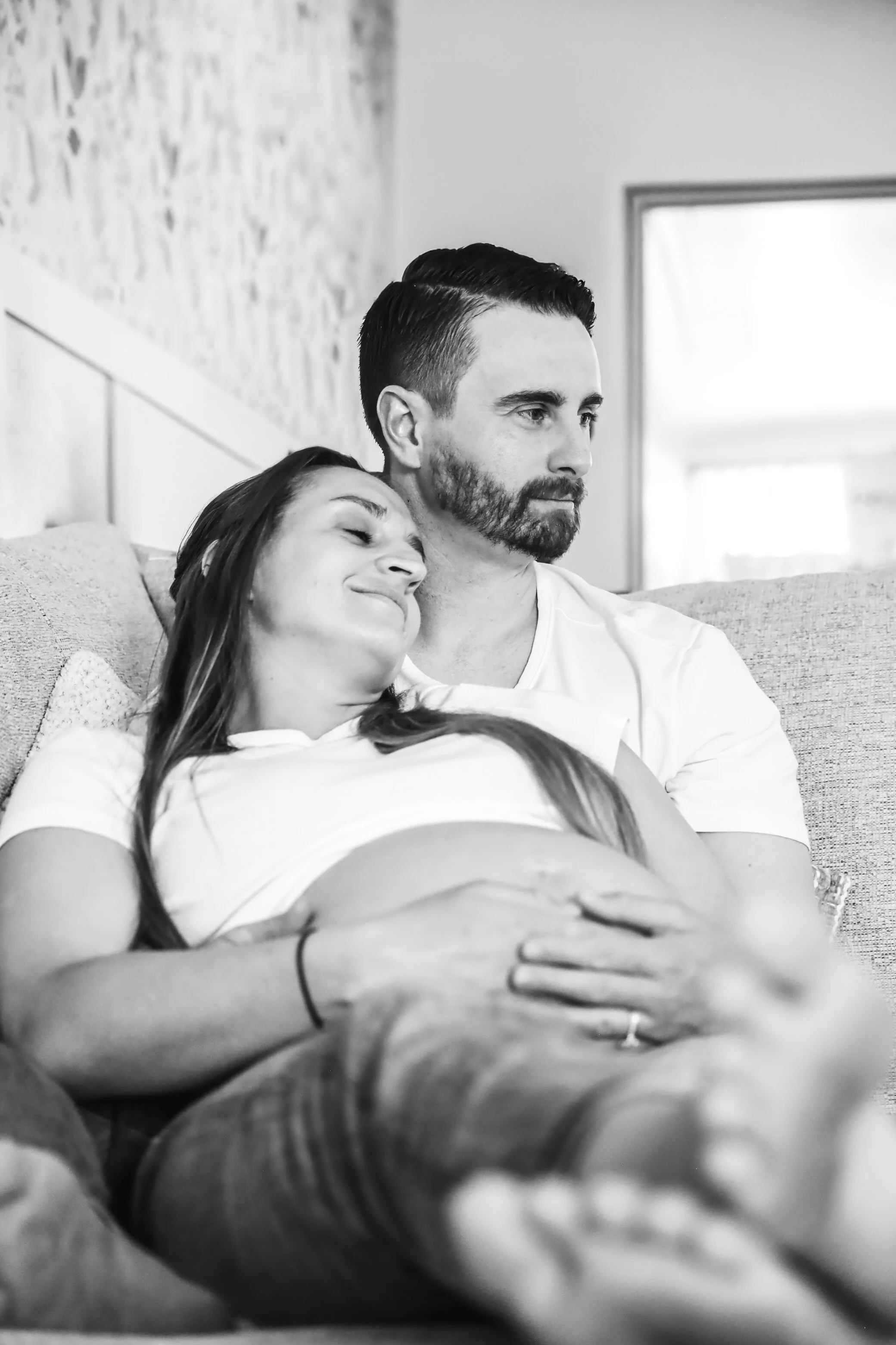 A black and white photo of a man and a woman sitting on a couch, with the woman resting her head on the man's shoulder. They appear happy and relaxed.