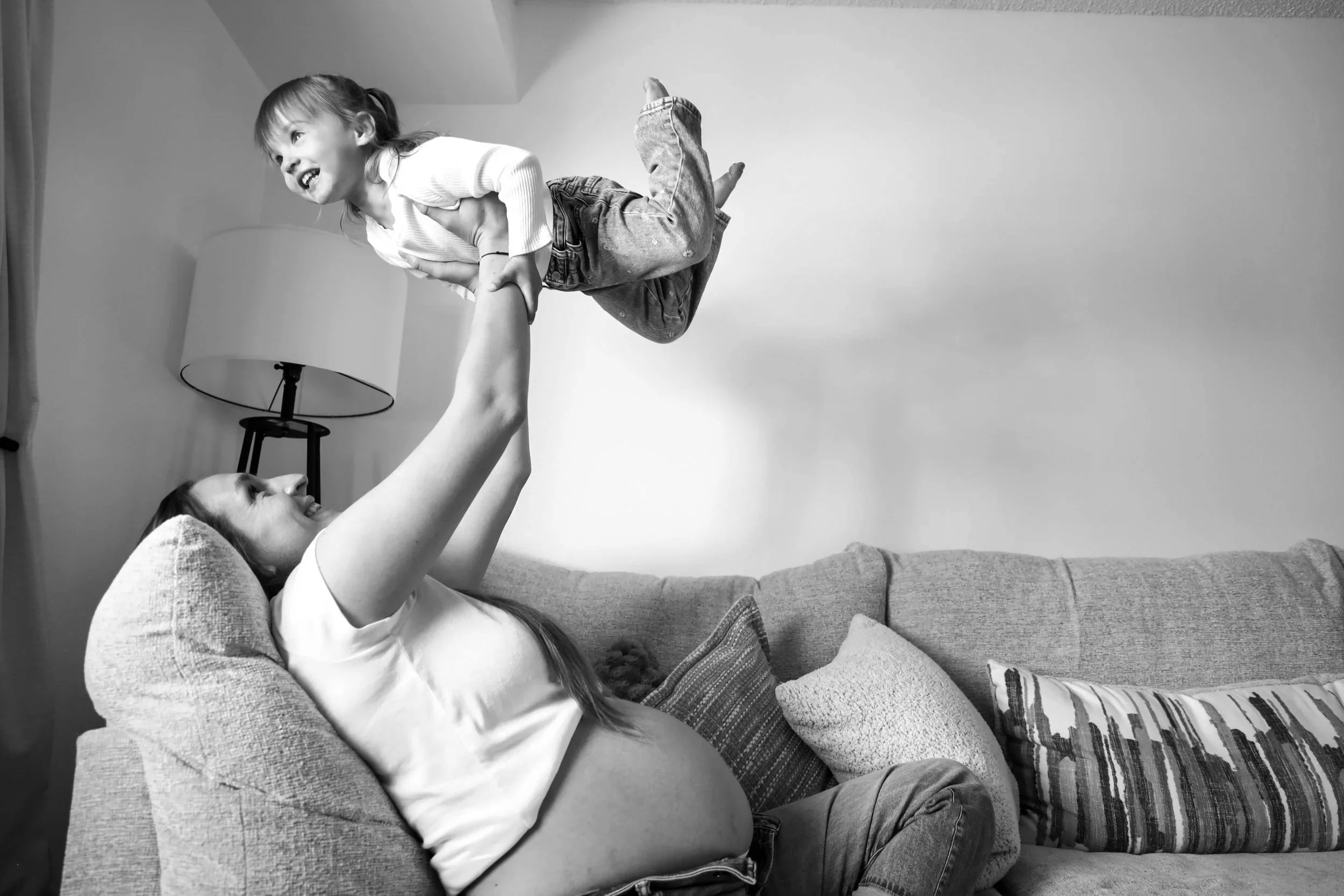 A woman with long hair and a white shirt is lying on a couch, lifting a young girl with pigtails and striped pants above her. The girl is smiling and wearing a long-sleeve shirt and jeans. A lamp and a curtain are visible in the background, and the entire scene is in black and white.