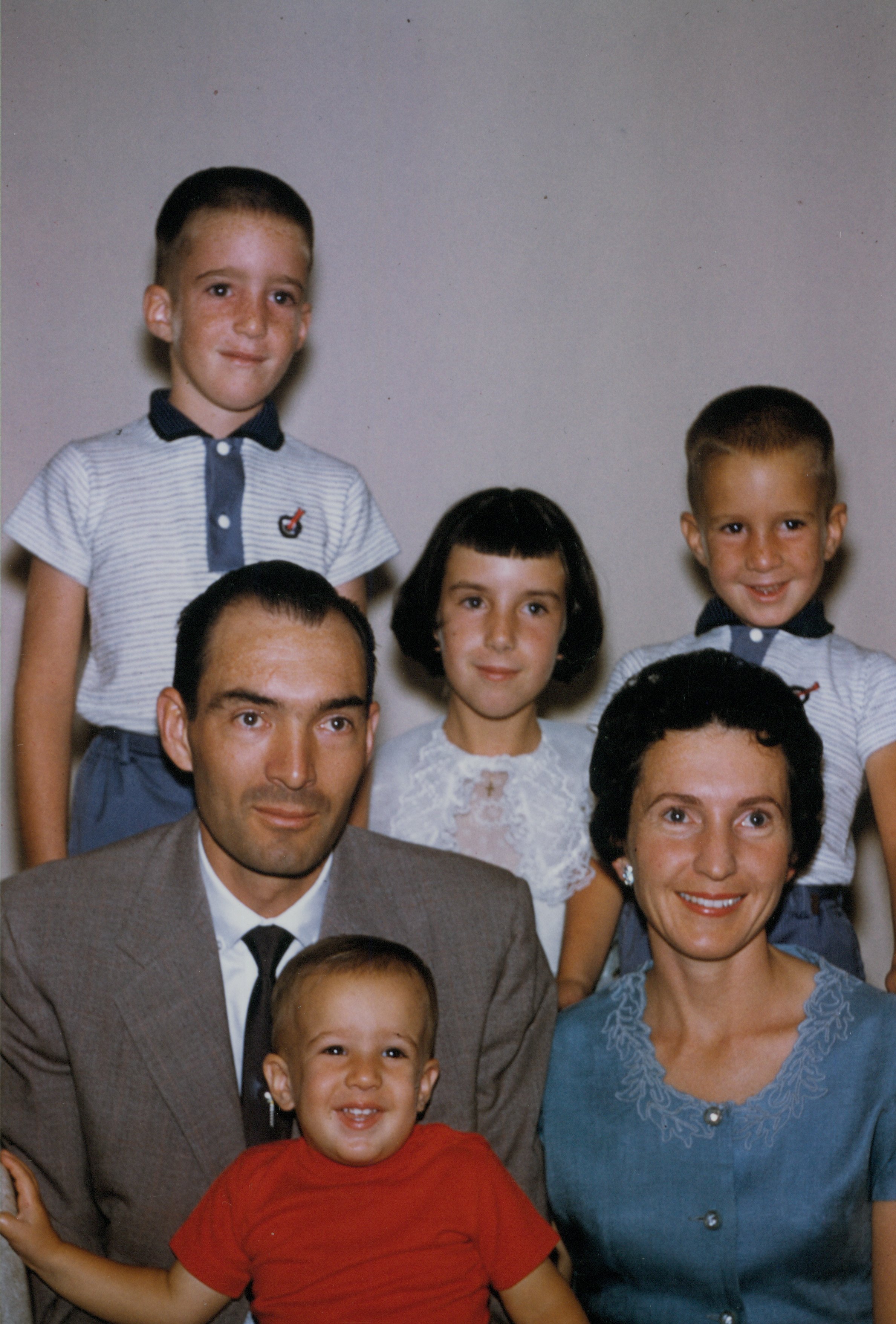 A group of children and adults posing for a family photo, with some children standing behind adults and a smiling young boy in a red shirt in the foreground.