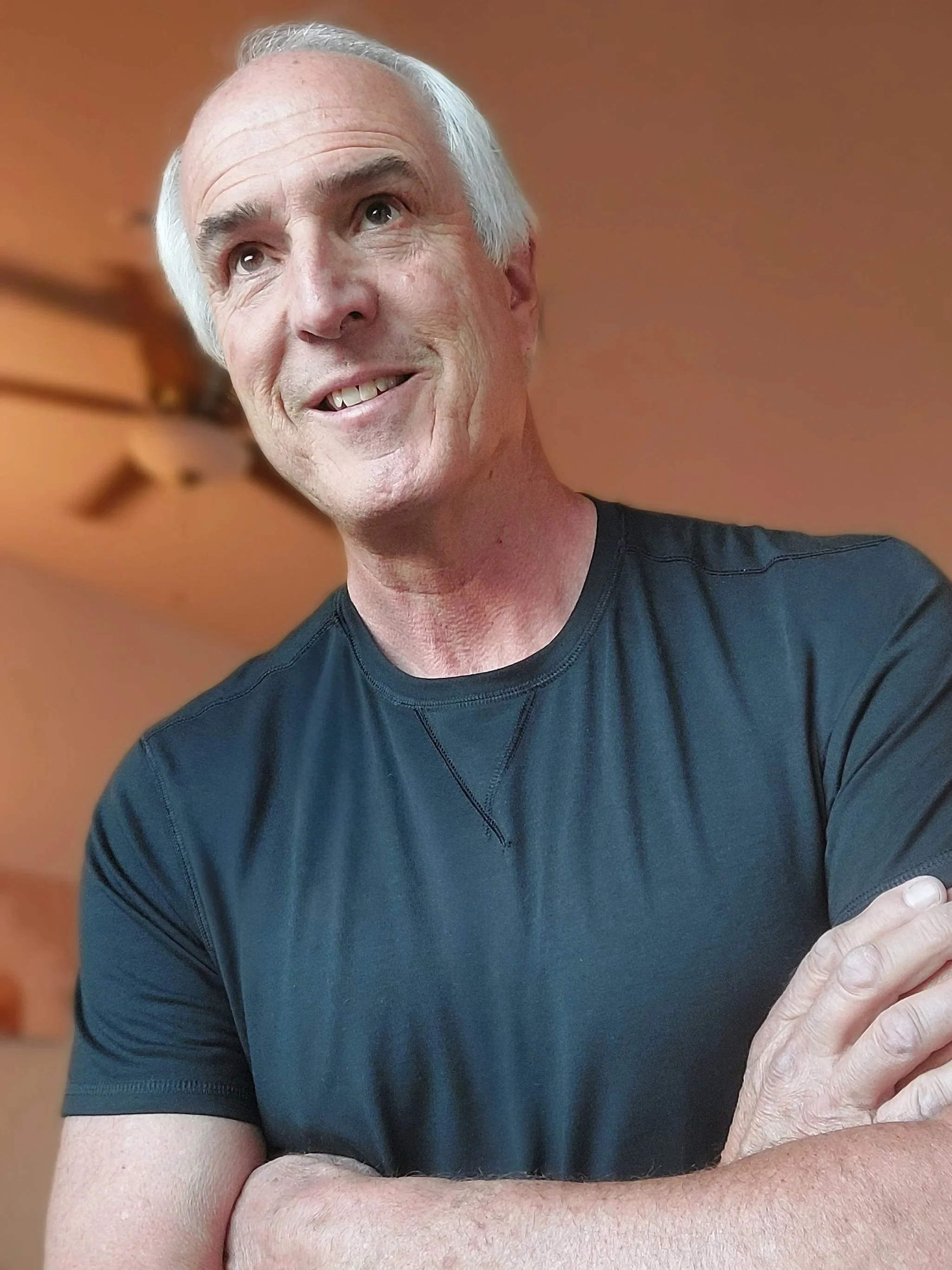 Close-up of an older man with white hair smiling, wearing a black shirt, showing his teeth, and resting his arms on a surface with his right hand visible in the frame.