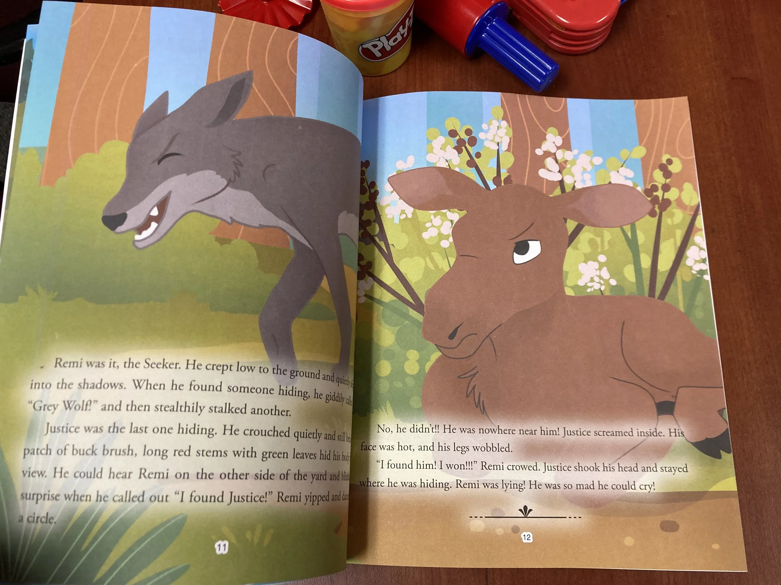 Open children's storybook showing illustrations of a grey wolf and a brown moose in a woodland scene with blooming bushes and trees.