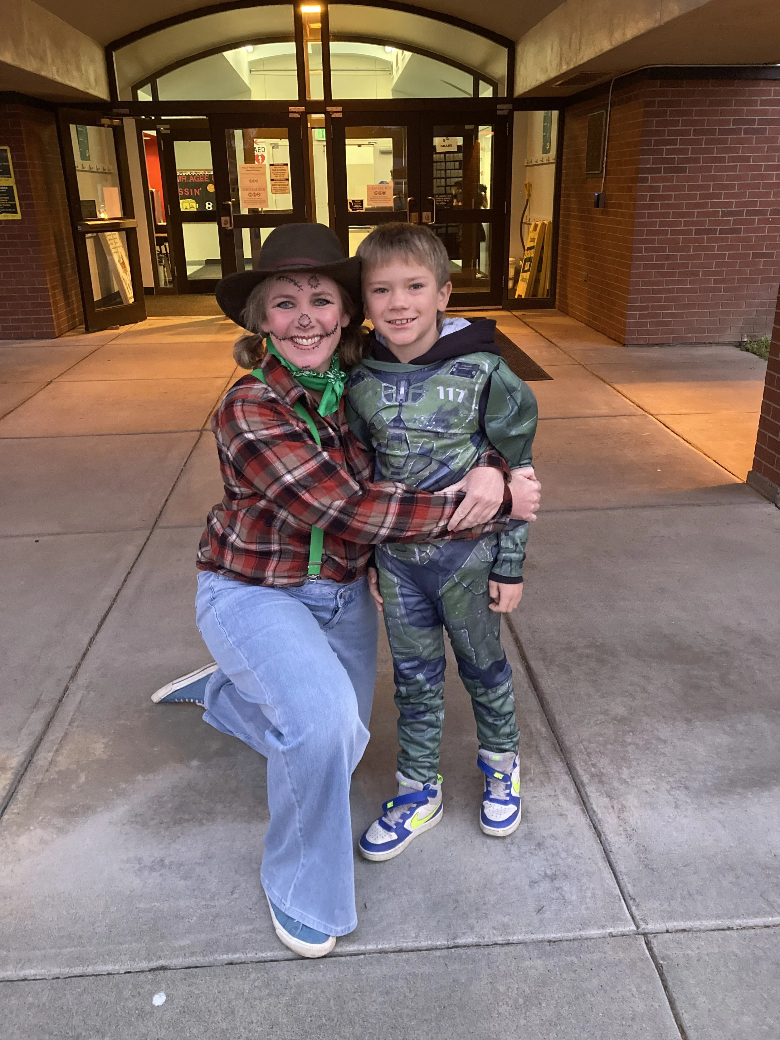 A woman with Halloween face paint and a hat, dressed in a plaid shirt and jeans, kneeling with a young boy in a green and black costume, outside a building at night.