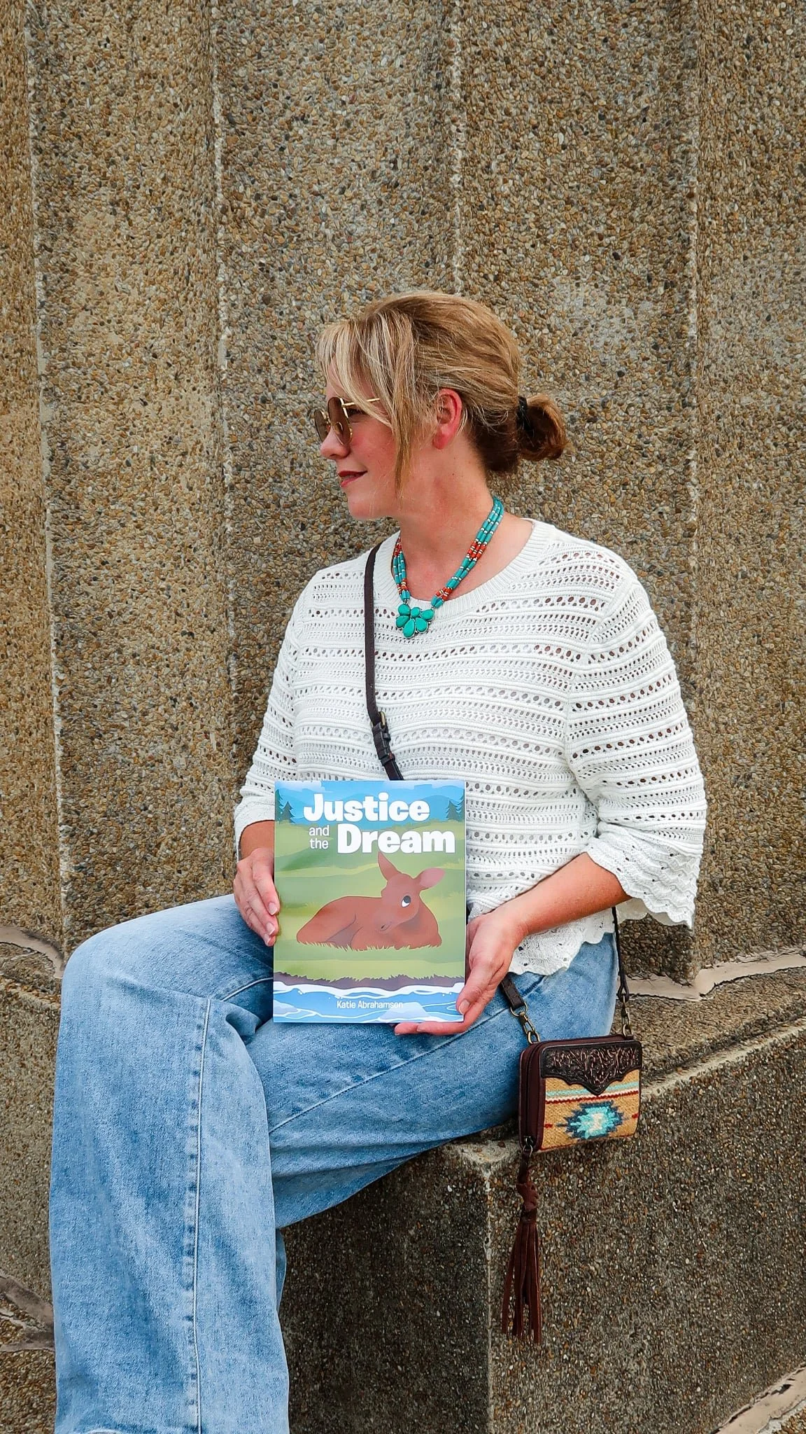 A woman in sunglasses and a white crochet top sitting against a stone wall, holding a children's book titled "Justice and the Dream" with an illustration of a young brown moose on the cover, with a small purse hanging from her shoulder.