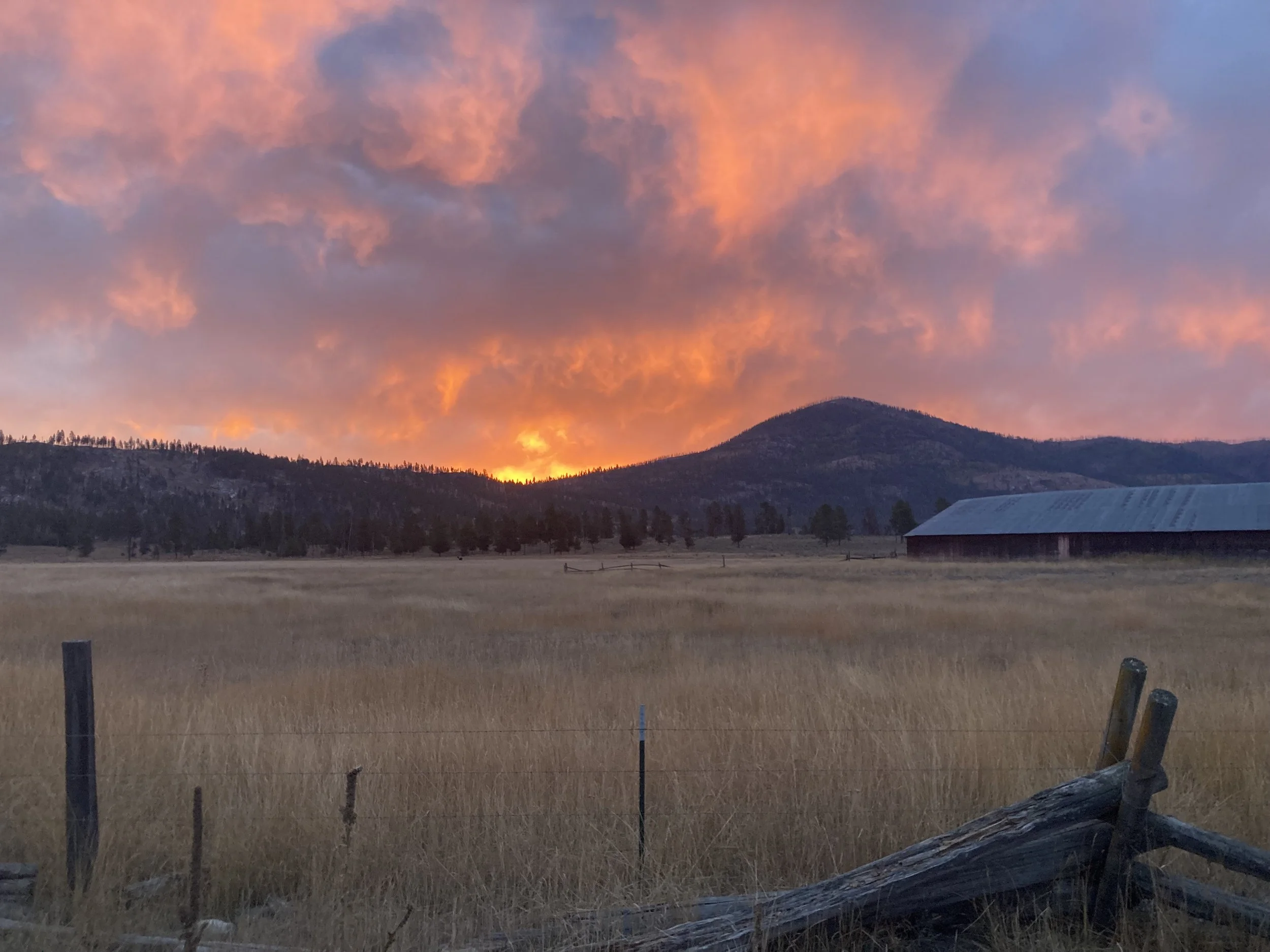 Sunset over mountains with orange and pink clouds in sky, rural field with a barn and fencing in the foreground.