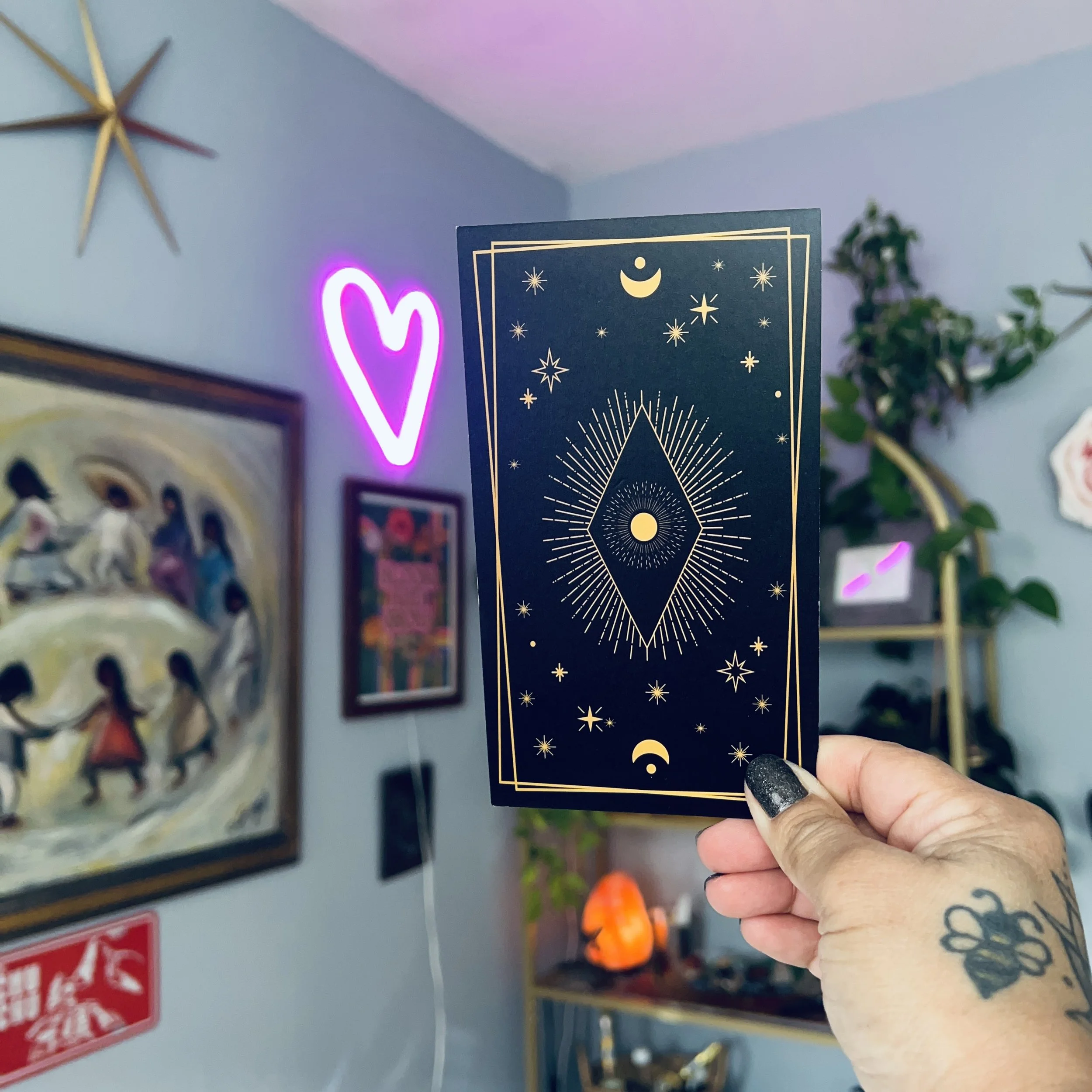 Person holding a tarot card with celestial imagery, stars, and a crescent moon, near a glowing pink heart-shaped neon sign.