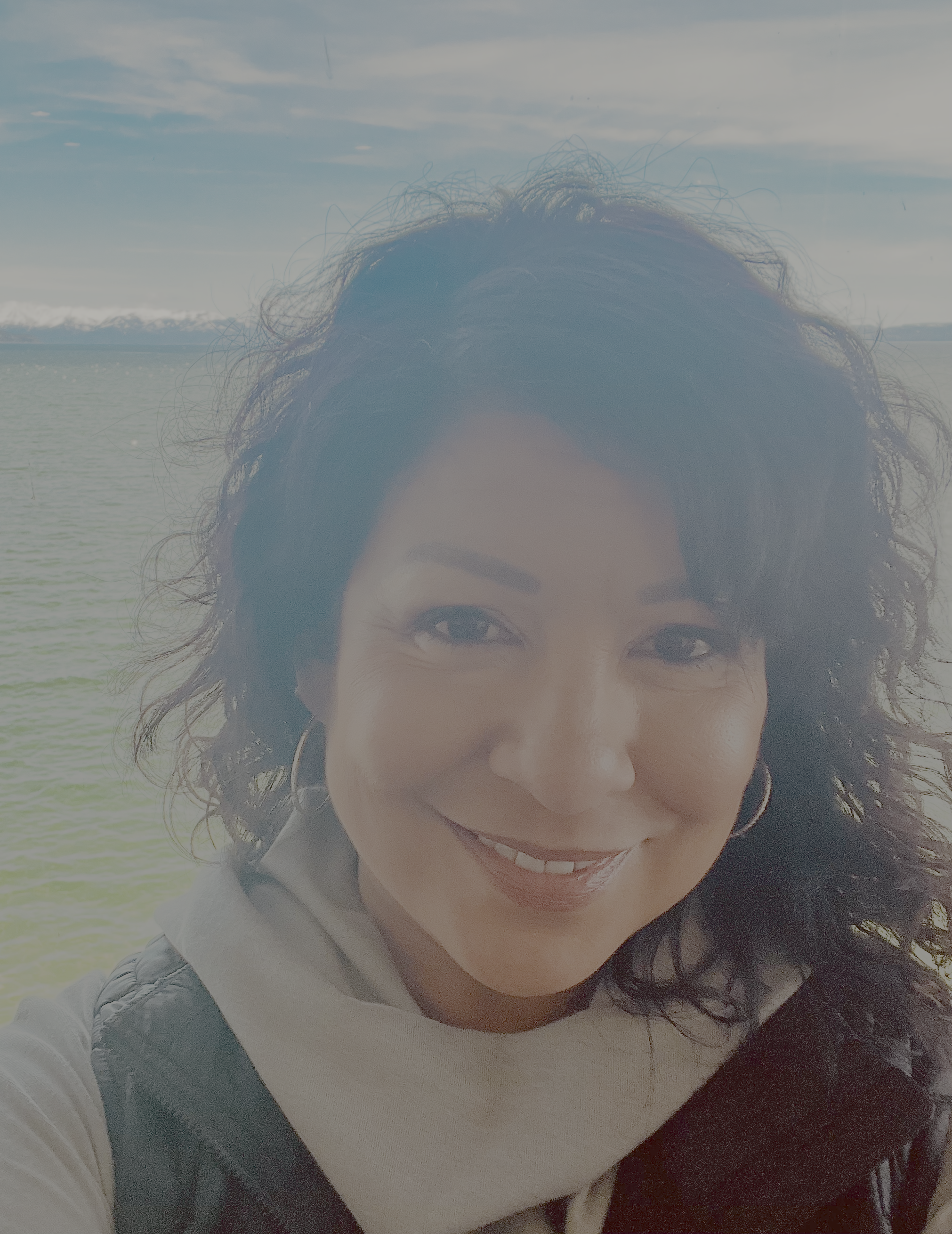 Close-up of a smiling woman with curly dark hair, wearing a white top and black vest, near a body of water with mountains in the background.