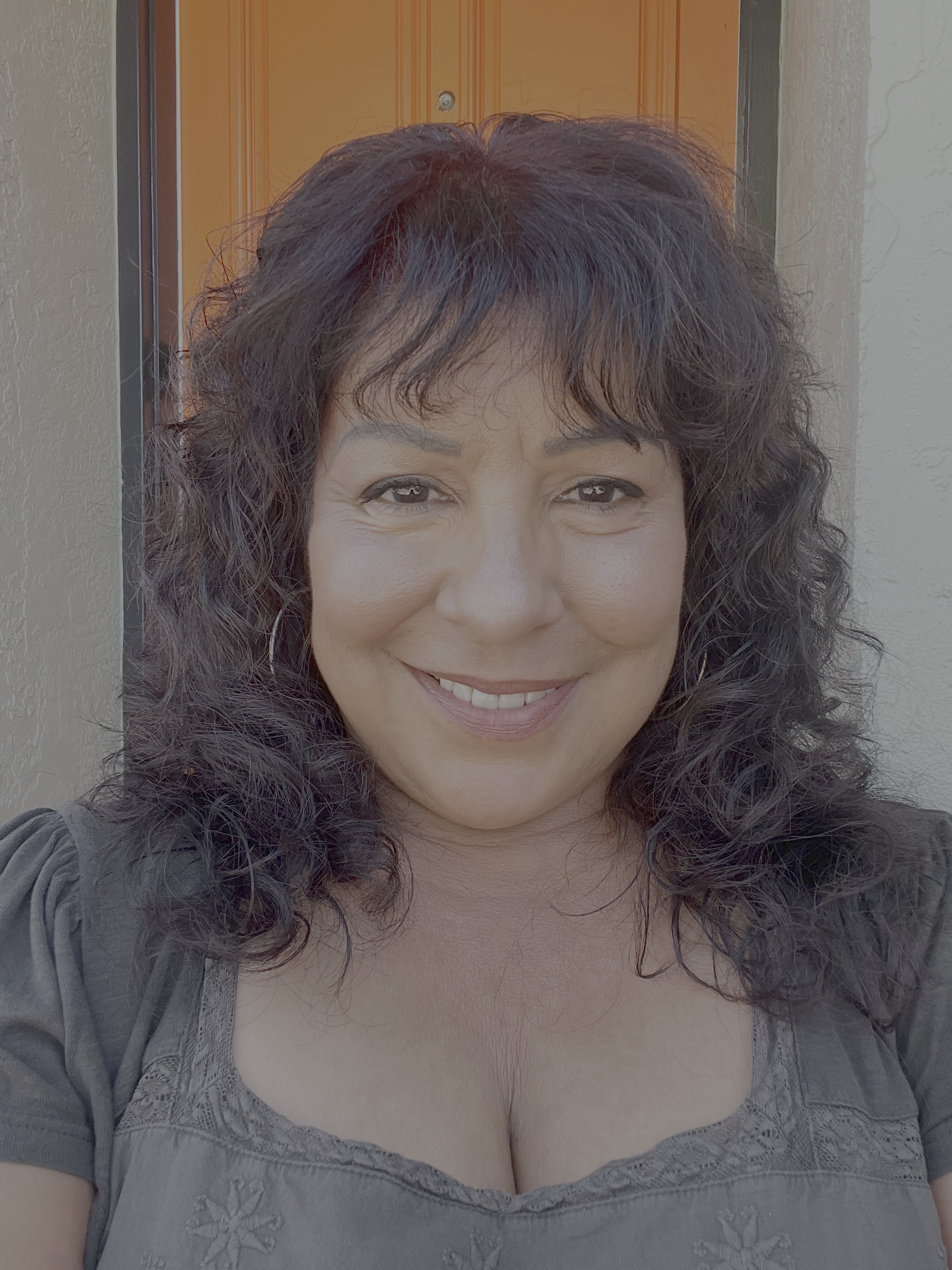 Close-up of a woman with dark, curly hair smiling, wearing a gray lace top, standing in front of a door and wall.