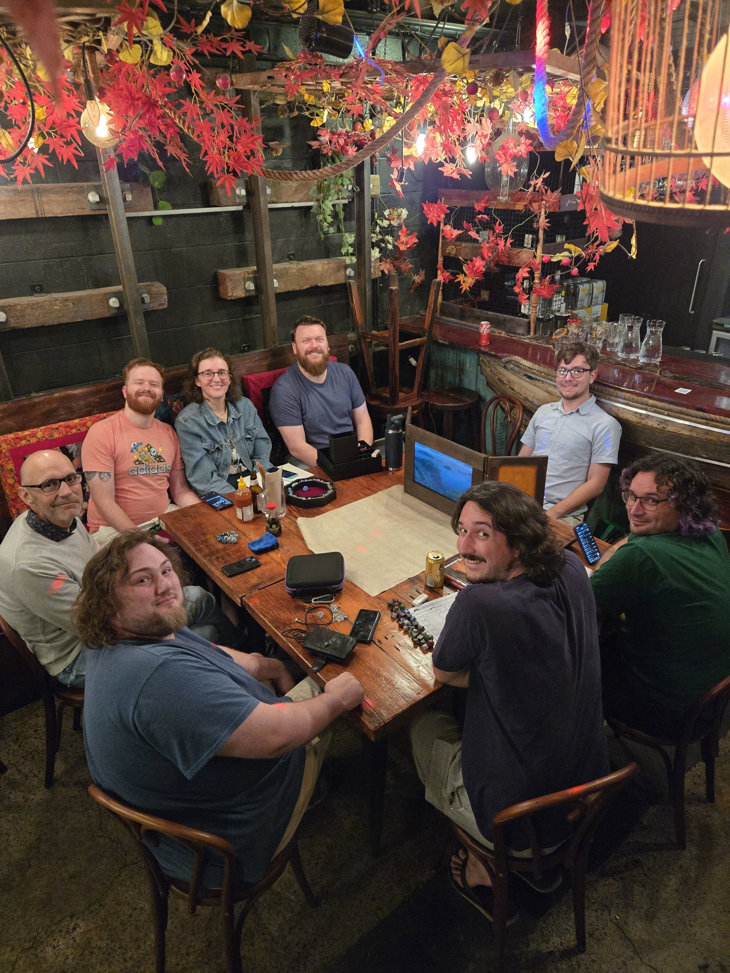 A group of people sitting around a wooden table in a cozy, decorated restaurant or cafe. The table has various gaming items, drinks, and snacks. The background features red autumn leaves and warm lighting, creating a welcoming atmosphere.