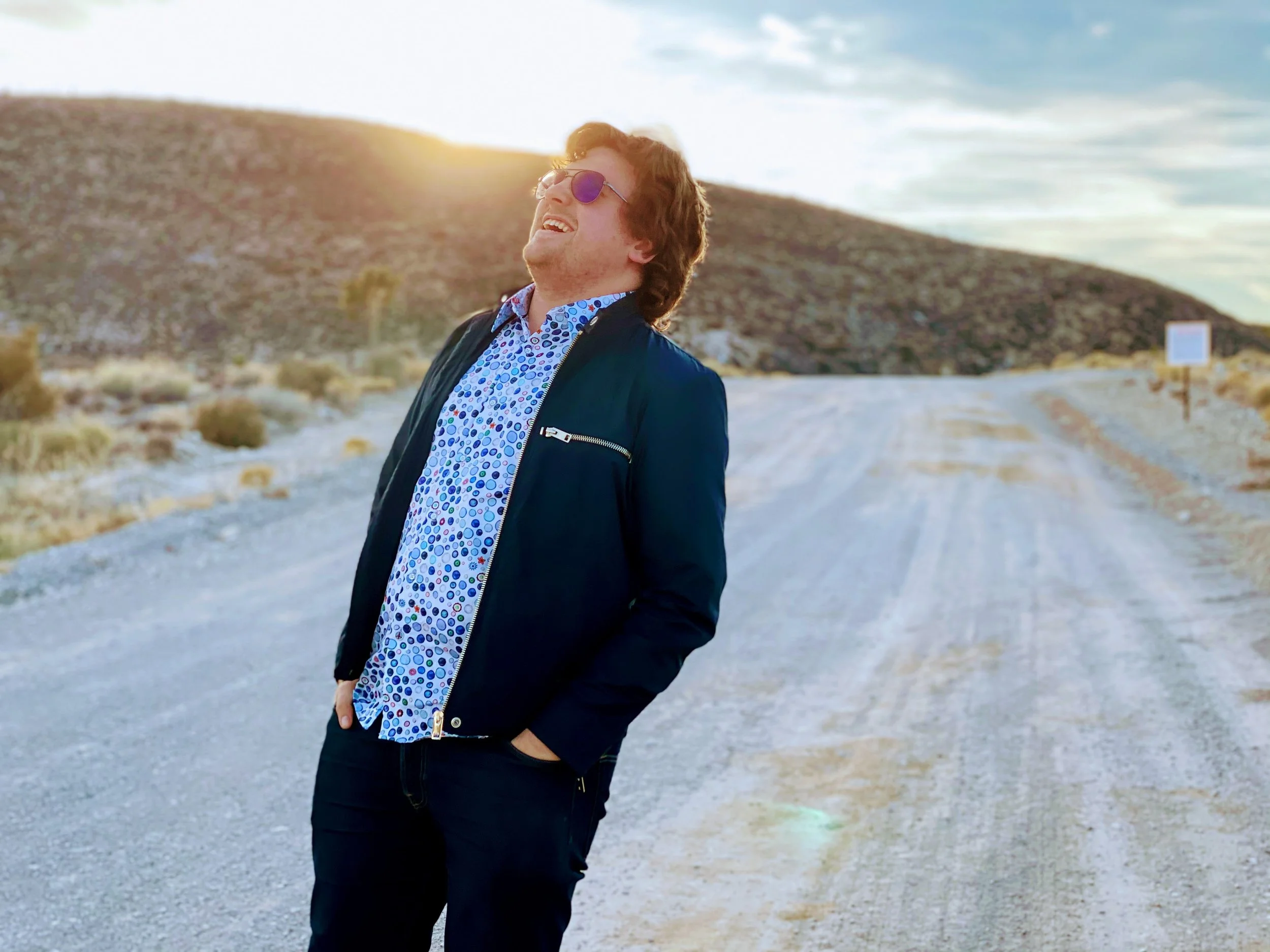A man with brown hair, wearing sunglasses, a patterned blue shirt, and a black jacket, stands on a dirt road with a hill and sky in the background, smiling and looking up towards the sun.