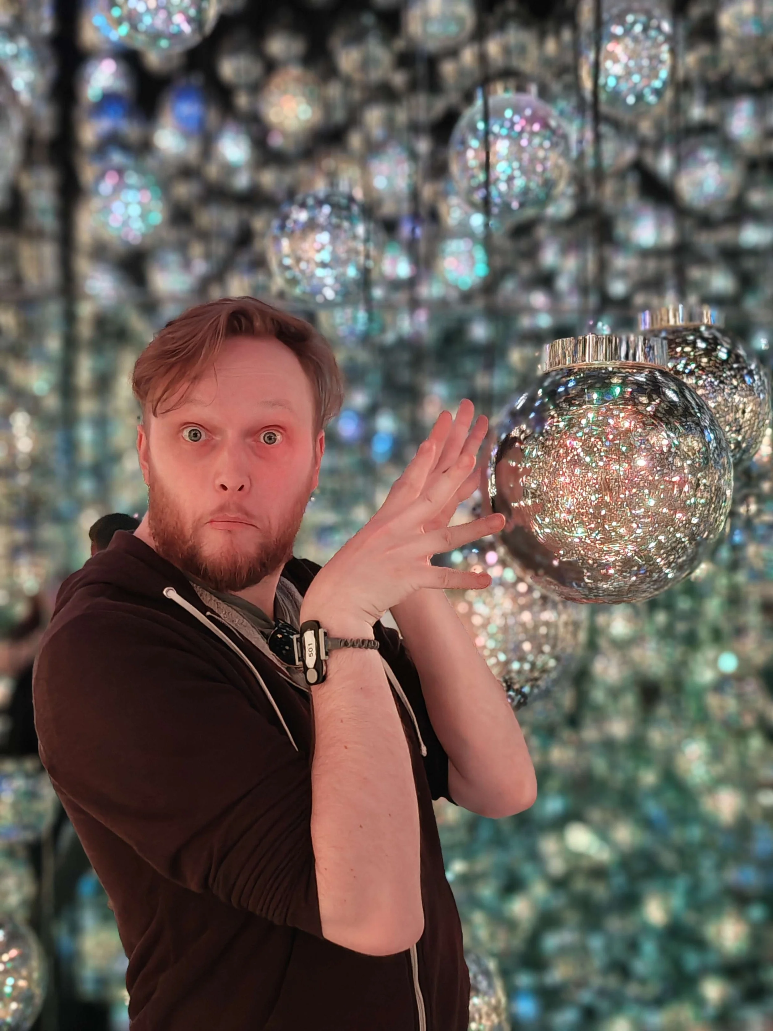 A man with a shocked expression, light brown hair, and a beard, holding a decorated Christmas ornament in front of a background filled with similar shiny ornaments hanging from the ceiling.