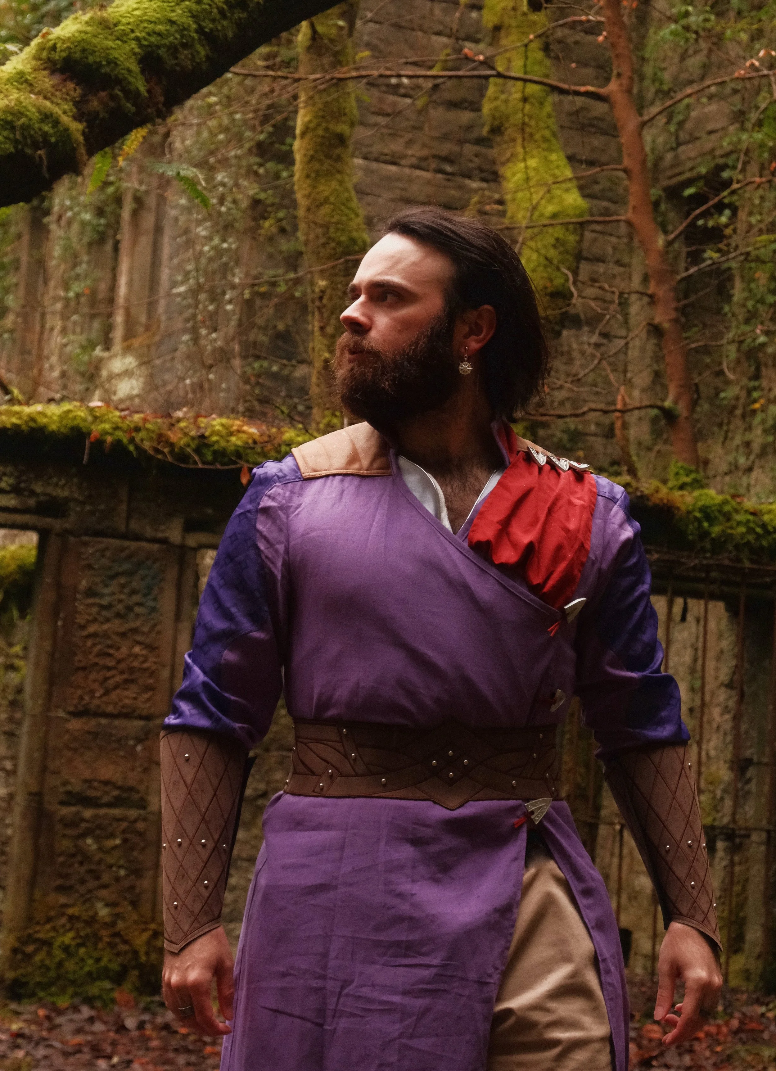 A man with long dark hair and a beard wearing a purple and red costume with fishnet sleeves stands outdoors in a forested area with moss-covered trees and a stone wall in the background.