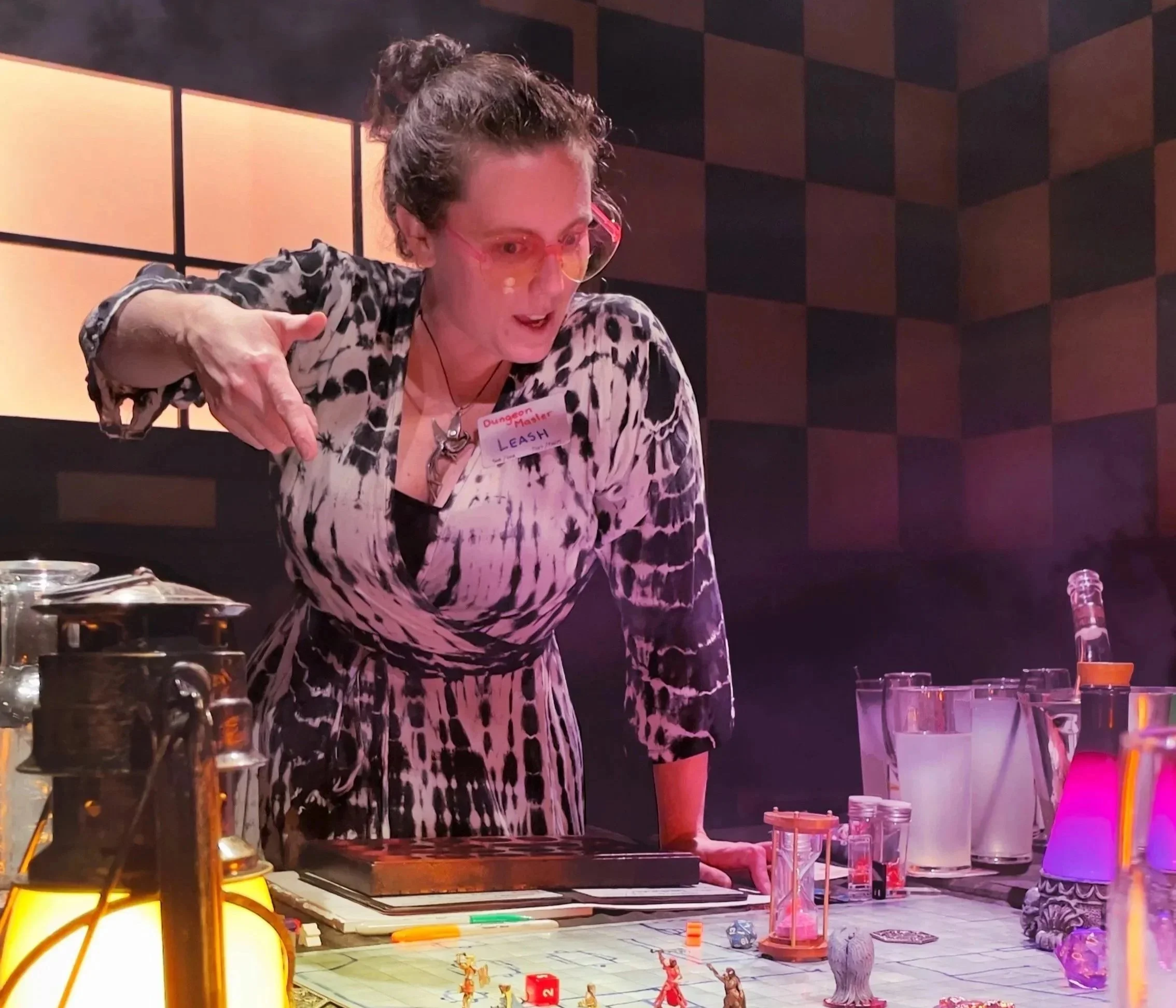 A woman wearing glasses and a patterned shirt is playing a tabletop role-playing game set up on a table with miniatures and dice. She has a name tag that reads "Dungeon Master Leash" and is pointing with her hand while speaking. The background features a dark, checkered wall with warm lighting.