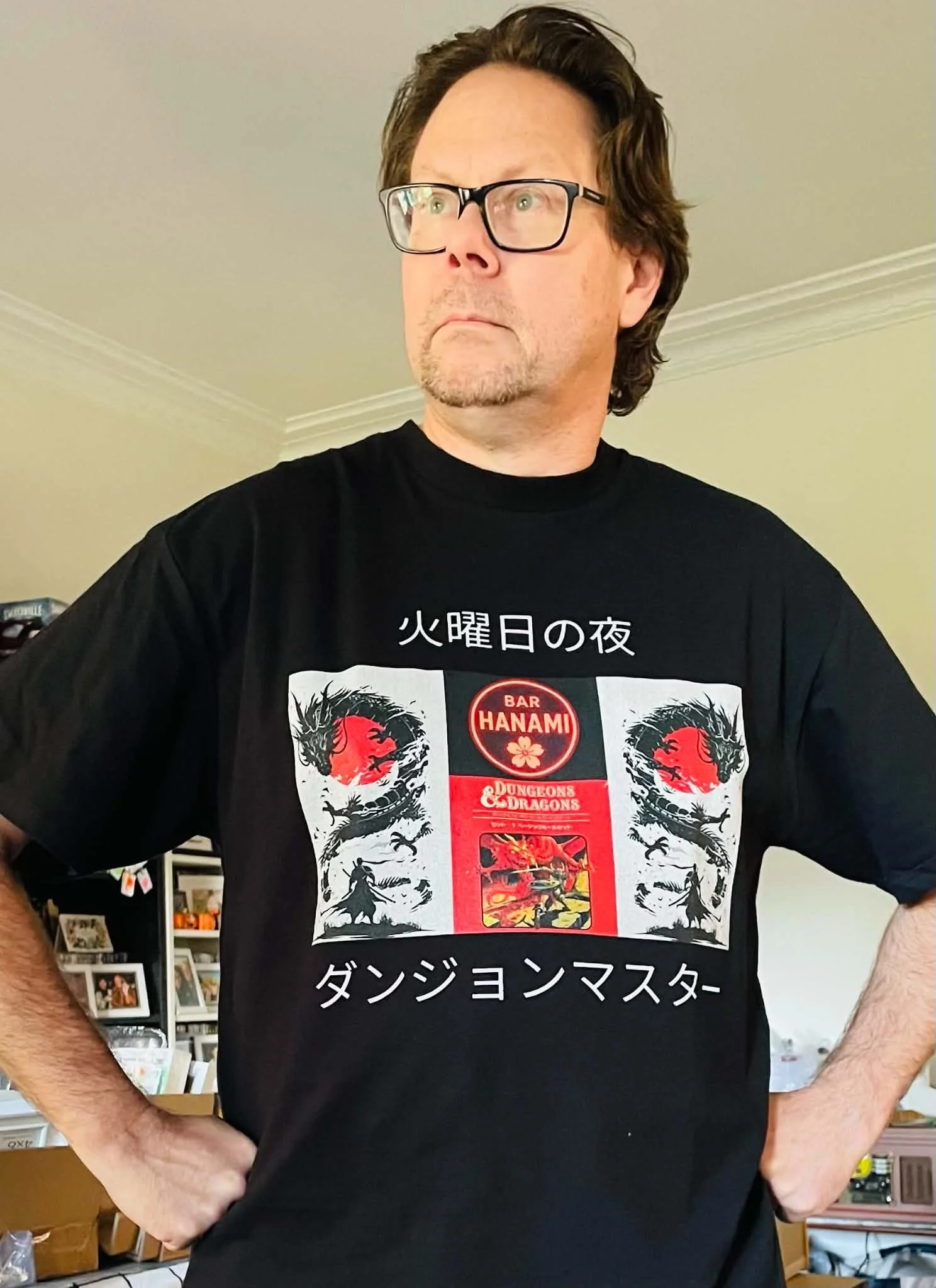 A man with brown hair, glasses, and a goatee wearing a black T-shirt with Japanese text and illustrations, standing indoors in front of bookshelves and frames.