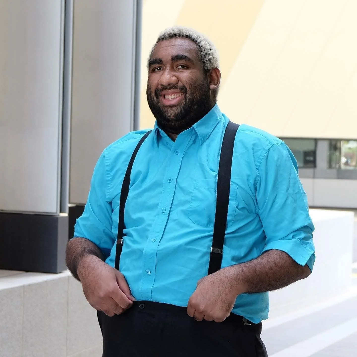 A smiling man with light gray hair and a beard, wearing a bright blue dress shirt with black suspenders, standing in an indoor setting with modern architecture.