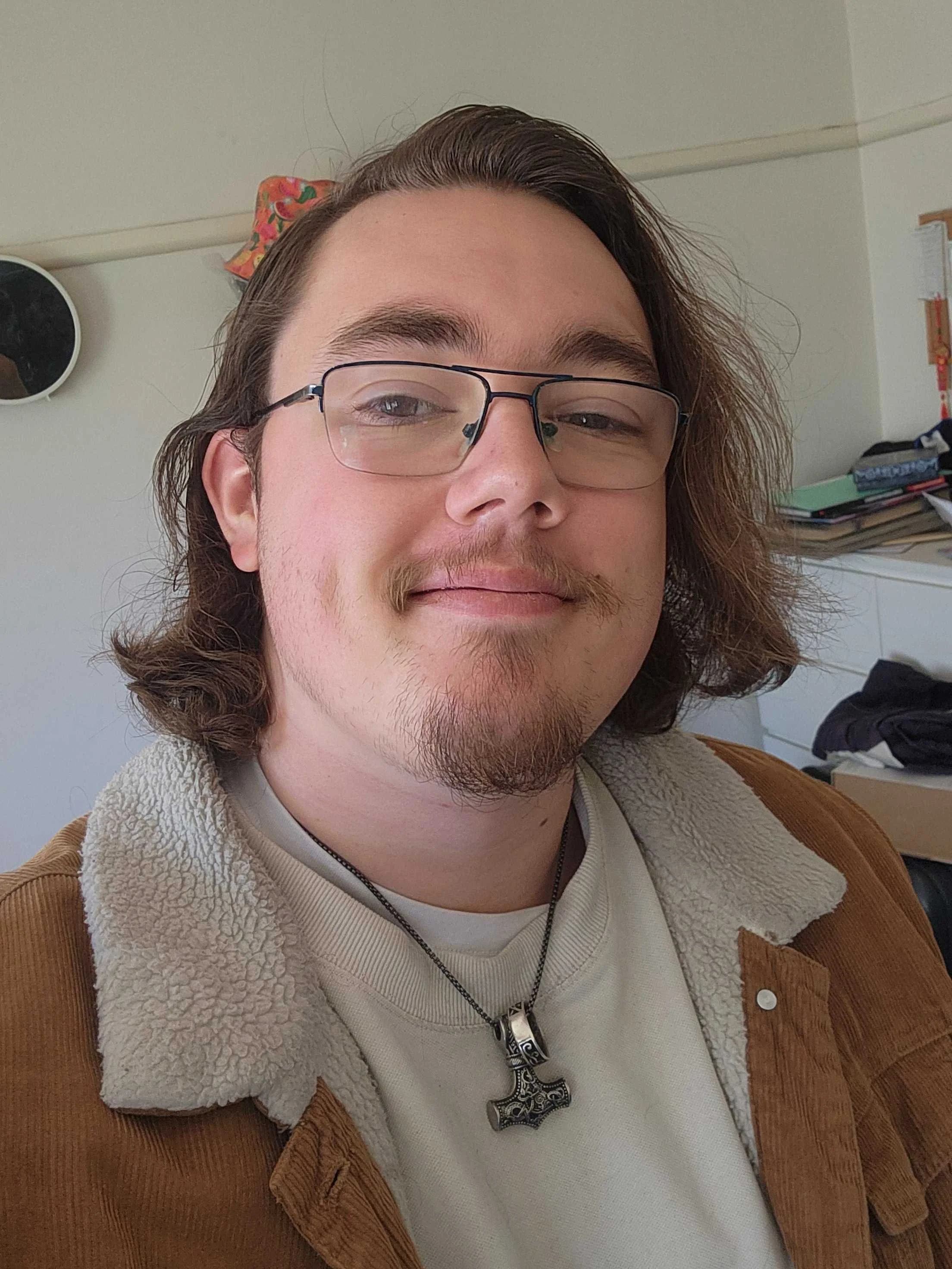 Close-up of a young man with long, wavy brown hair, wearing glasses, a beige sweater, and a brown suede jacket with a fleece collar, sitting indoors with a background of shelves and books.