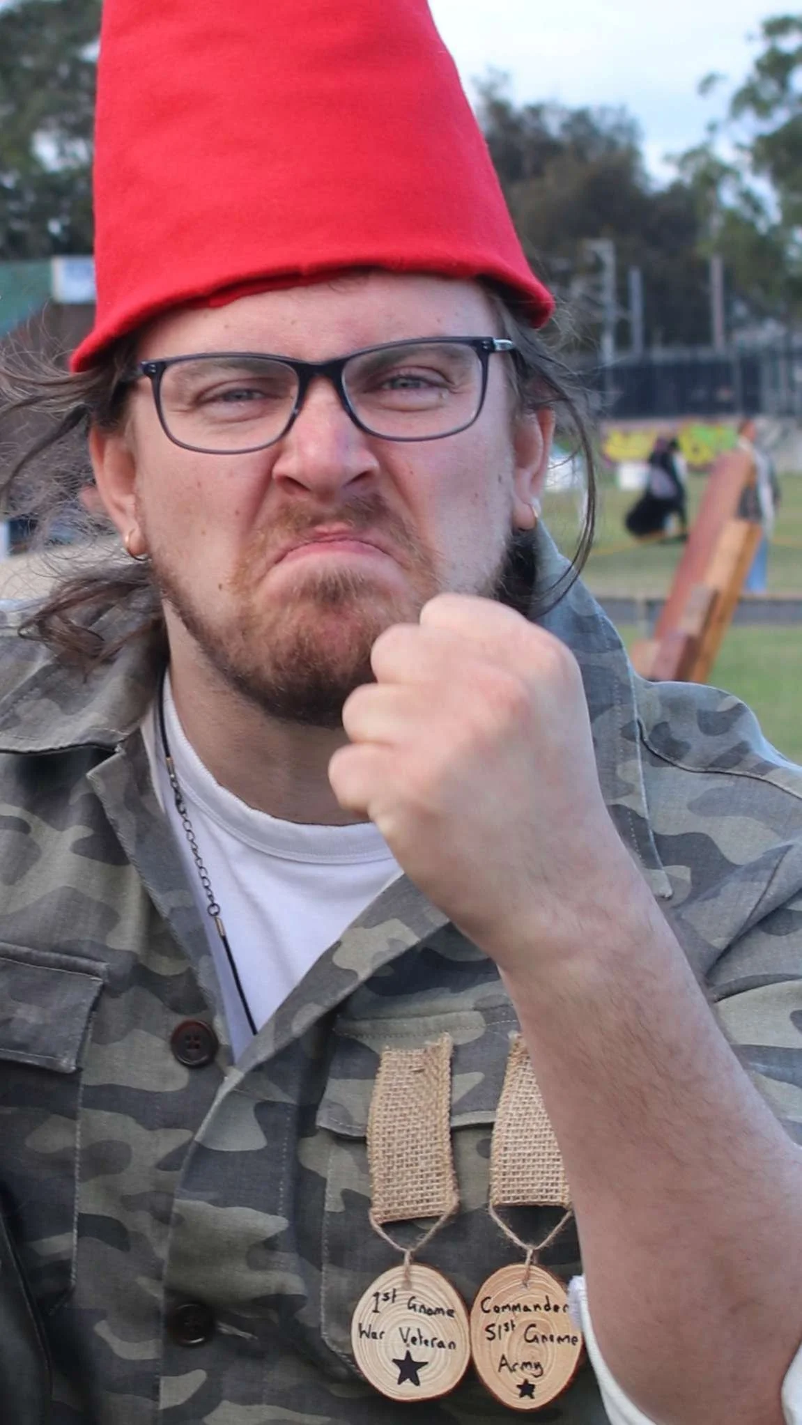 A man wearing glasses, a red hat, and camouflage jacket with military medals around his neck, standing outdoors. In the background, people are at a park or outdoor event.