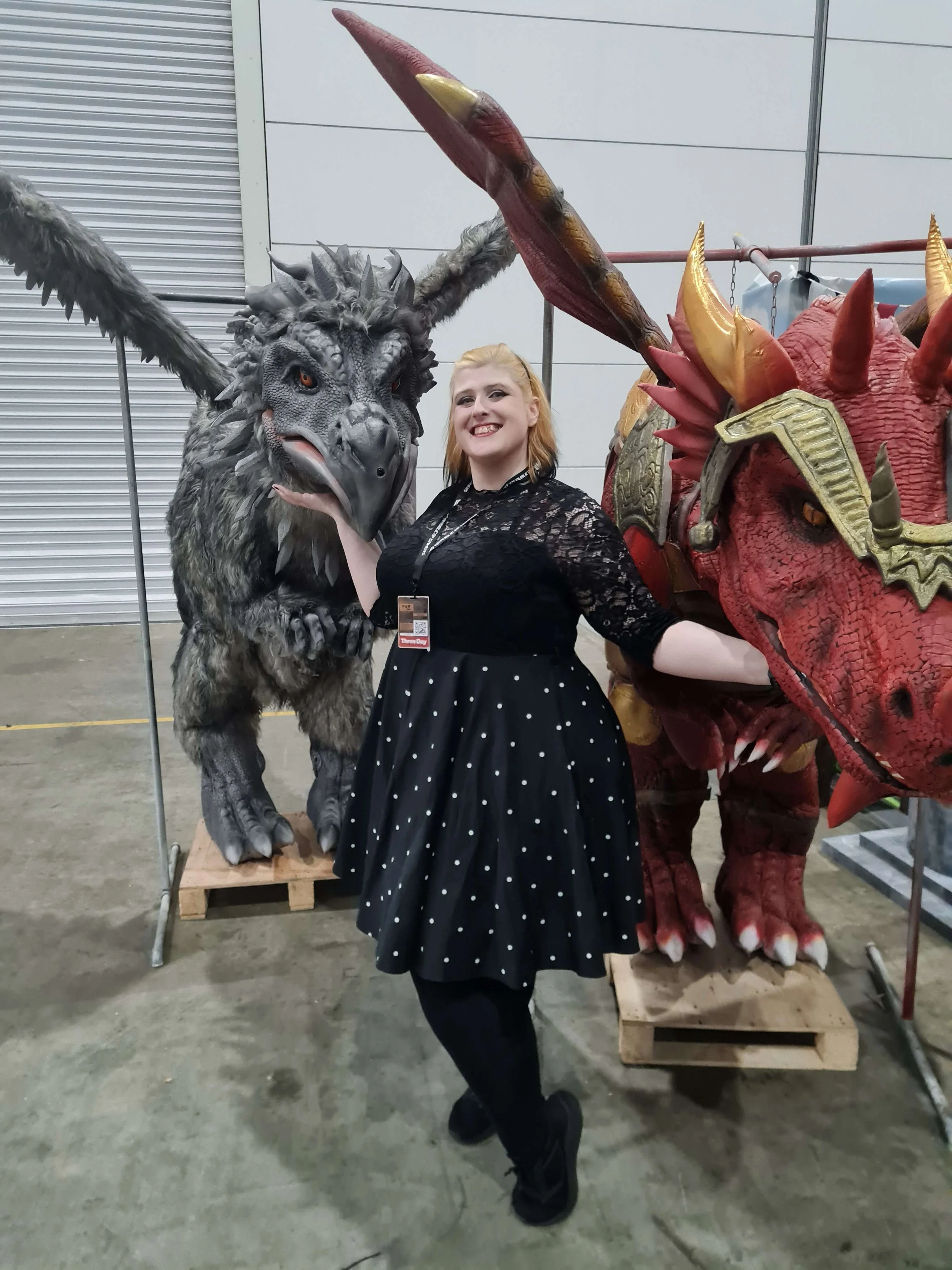 A woman with blonde hair in a black dress with white polka dots is smiling and posing between two large costume creature models, one resembling a horned dragon and the other a winged beast, at an indoor event.