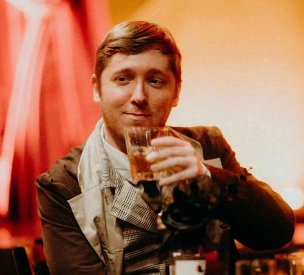 A person with short brown hair and a beard holding a glass of whiskey in a dimly lit room with reddish-orange curtains in the background.