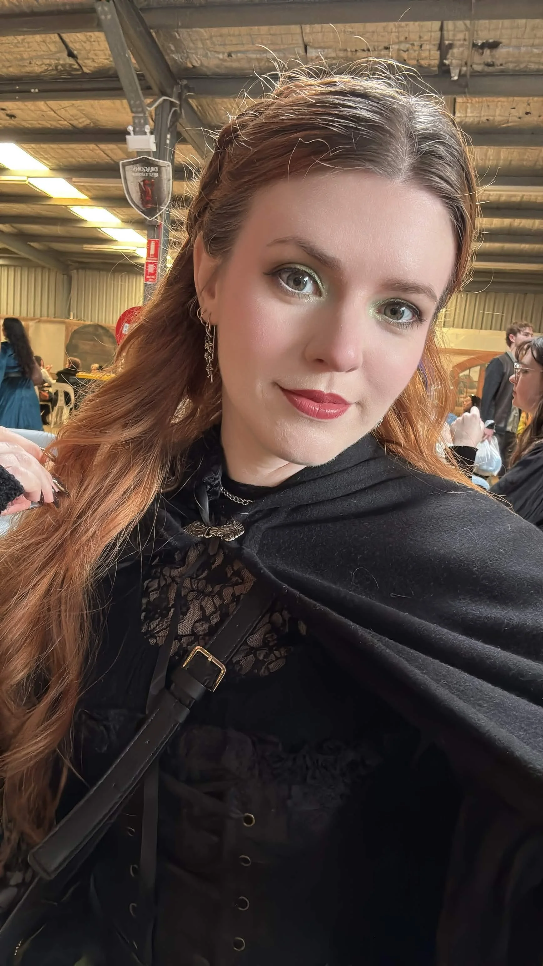 A young woman with light skin, long reddish-brown hair, and eye makeup, sitting in what appears to be a large indoor venue with a metal ceiling and other people in the background.
