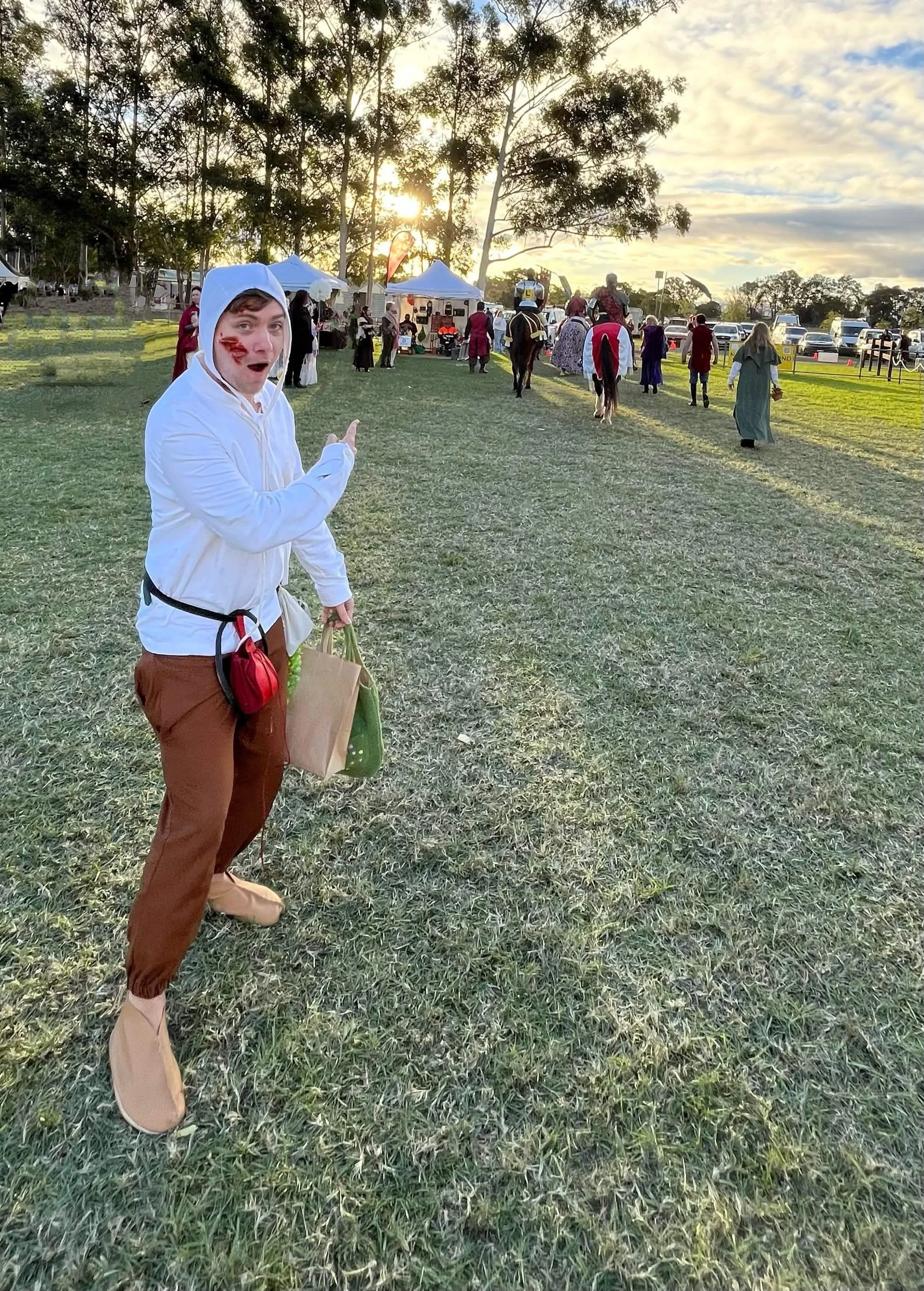 A person dressed as a movie character with face paint, pointing at the camera, at an outdoor event with tents, people in costumes, and trees in the background during sunset.