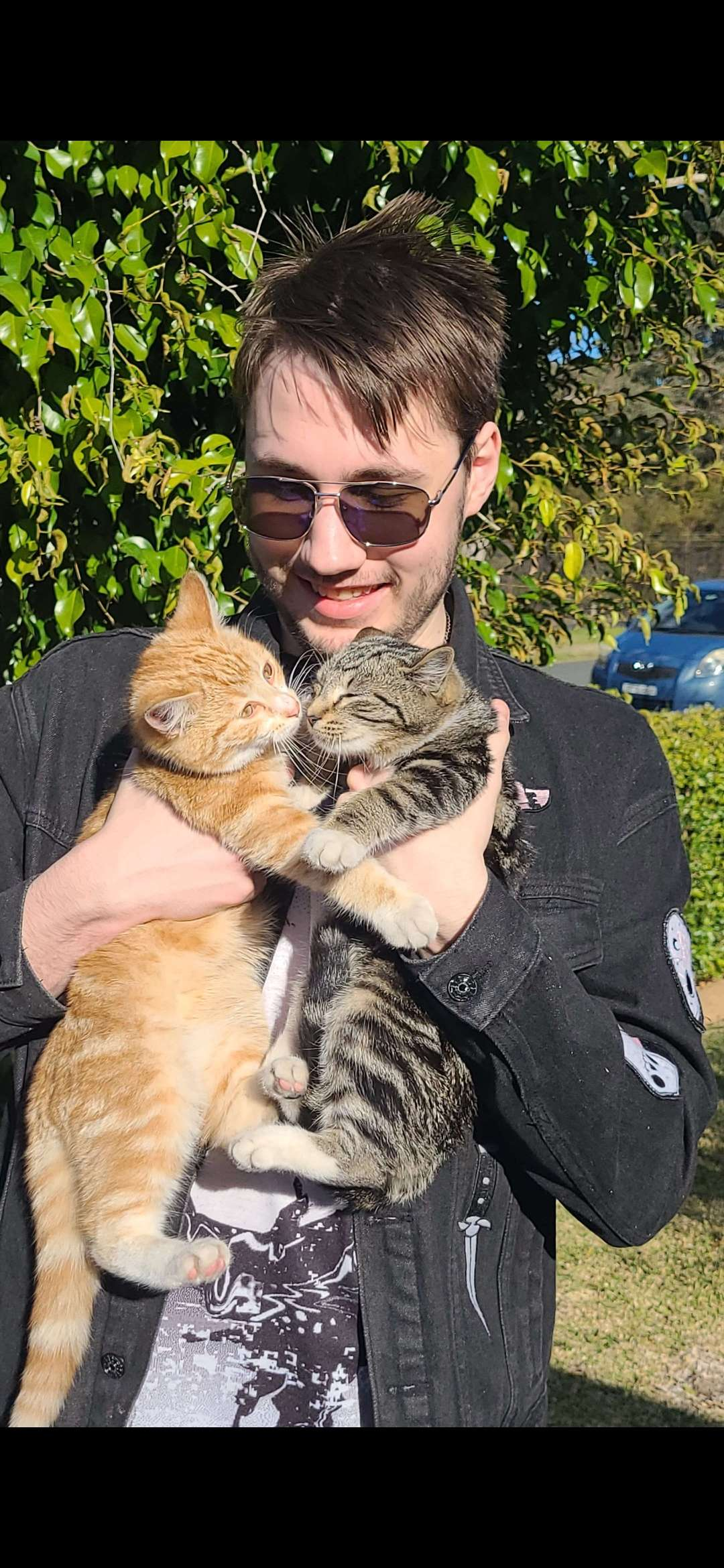 A young man with sunglasses holding two cats, one orange tabby and one gray tabby, outside in front of green leafy bushes and a parked car.
