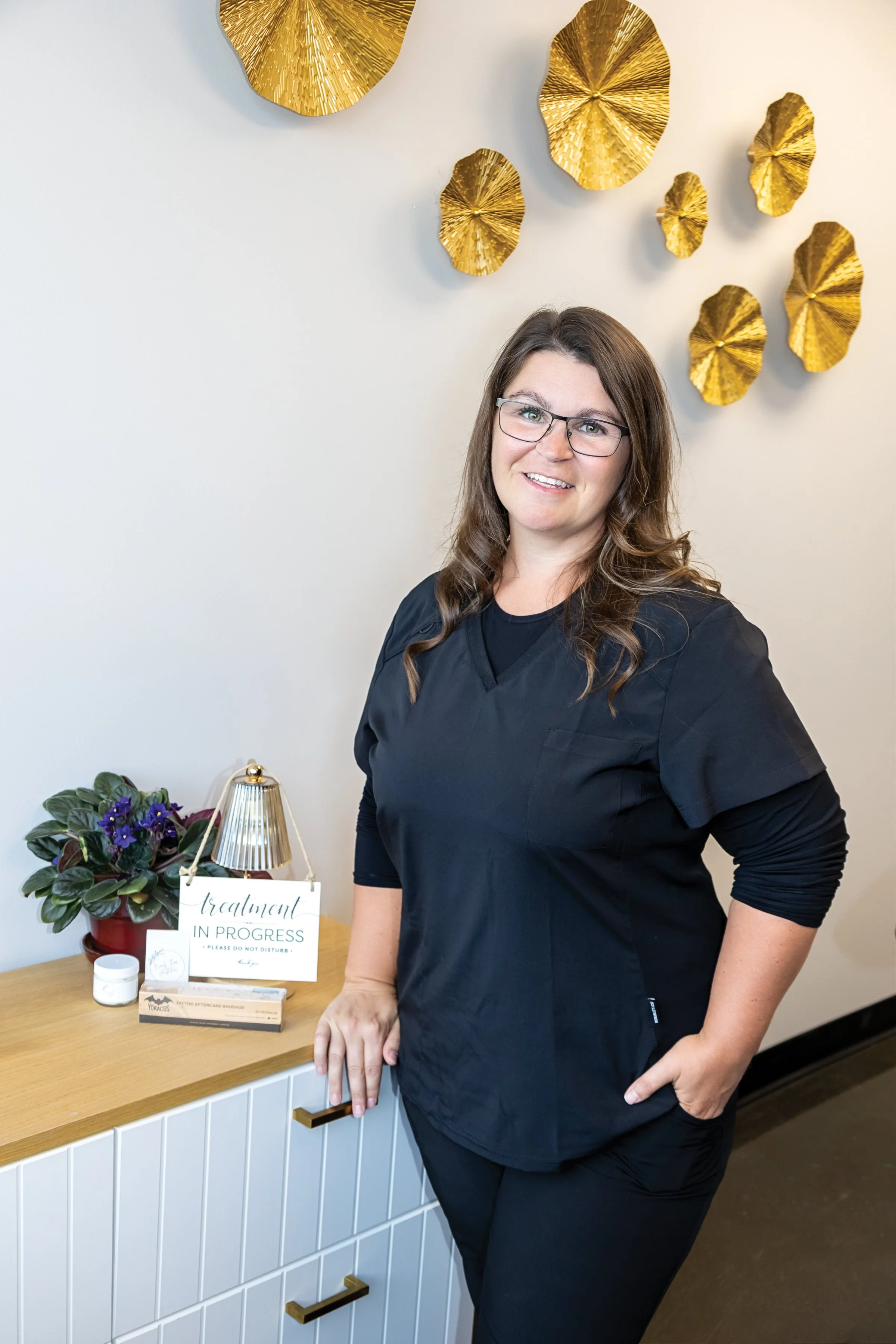 A woman dressed in black medical scrubs standing next to a wooden table with a potted plant, a sign that says 'treatments in progress, please do not disturb,' and some small items. She is smiling and wearing glasses, with a decorative wall hanging of gold-colored leaves behind her.