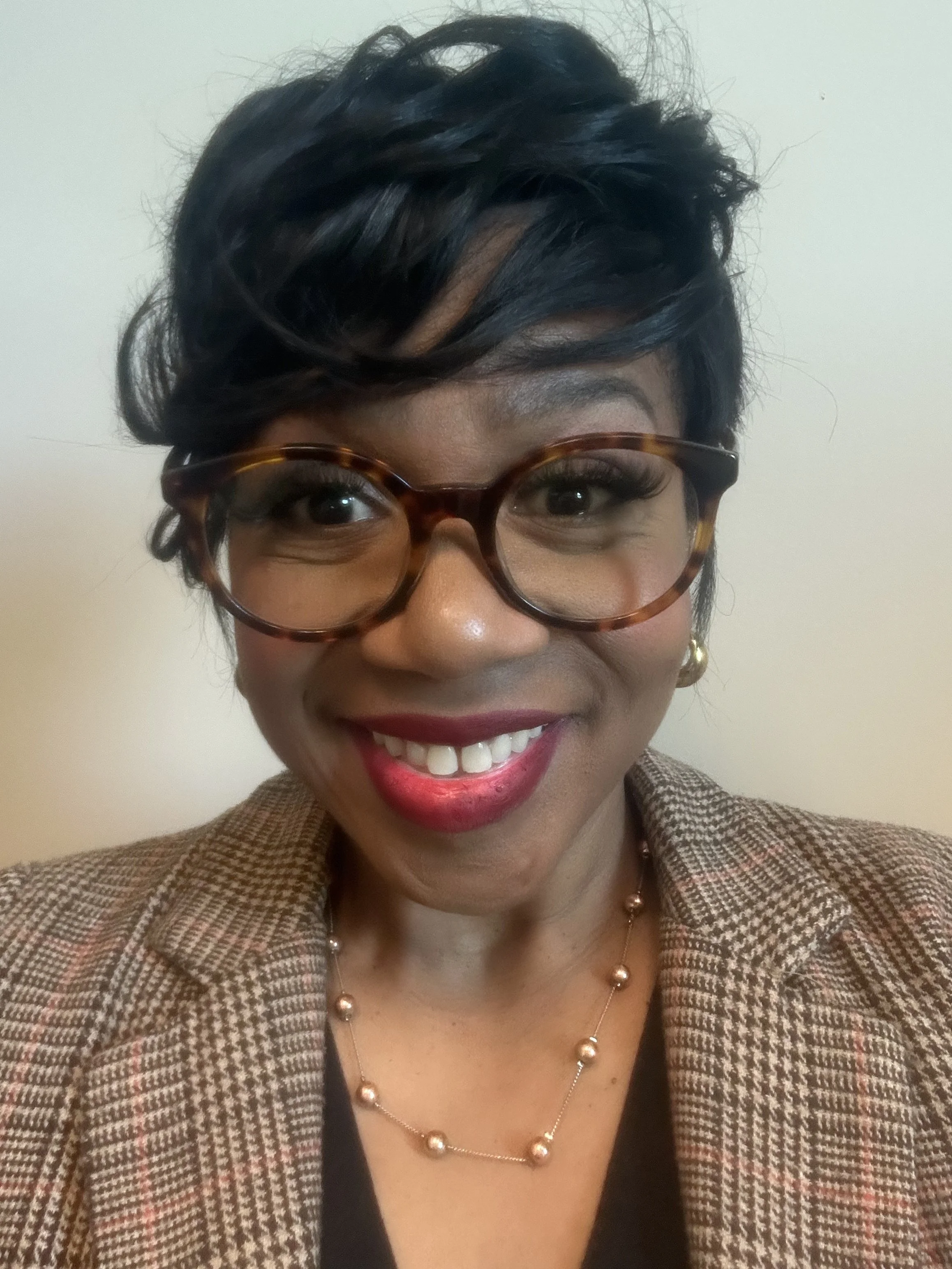A smiling woman with short black hair, wearing large tortoiseshell glasses, a checkered blazer, a black top, and a gold pearl necklace in front of a plain white wall.