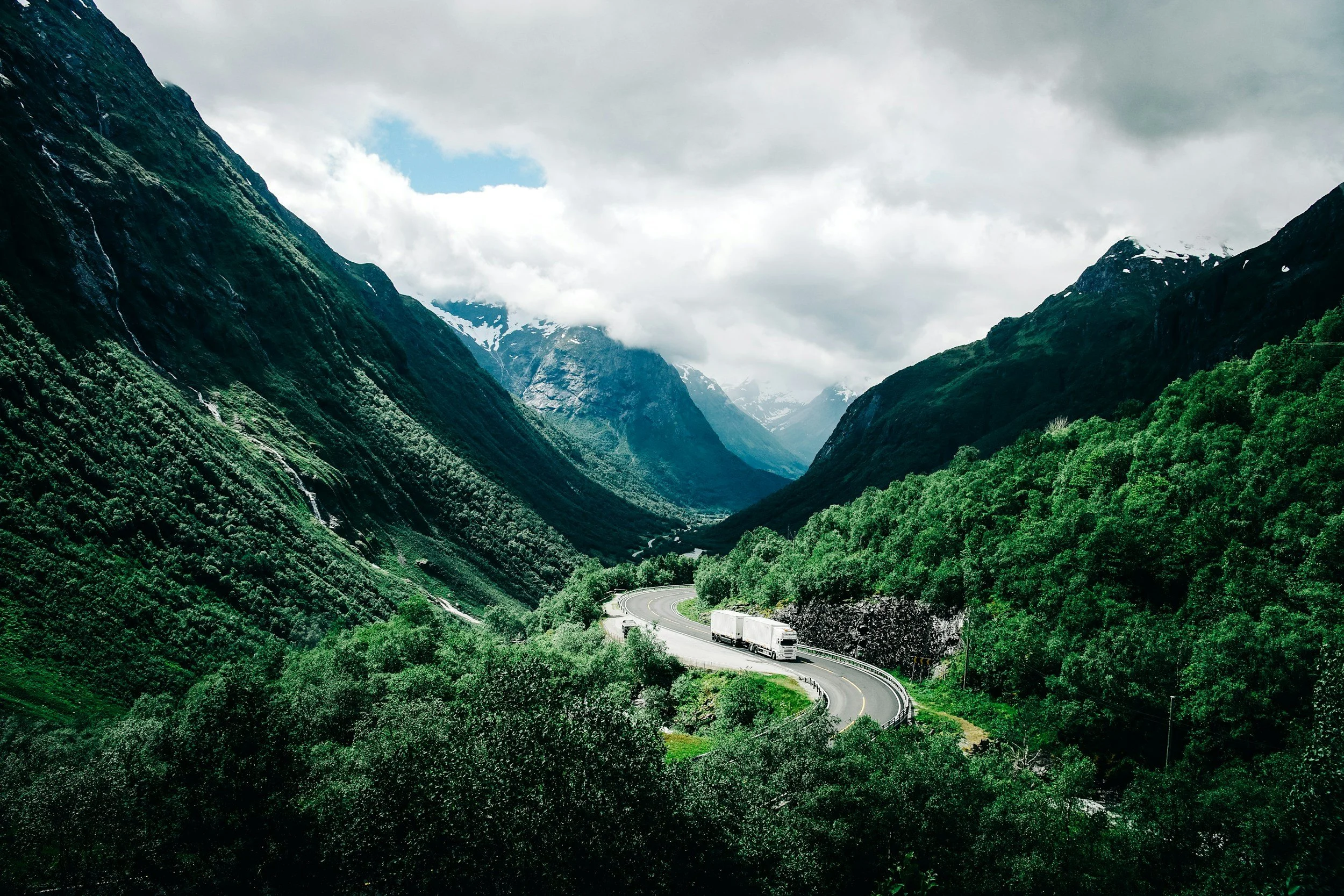 A winding road runs through a green valley surrounded by tall, forested mountains with some snow near the peaks, under a cloudy sky with patches of blue.