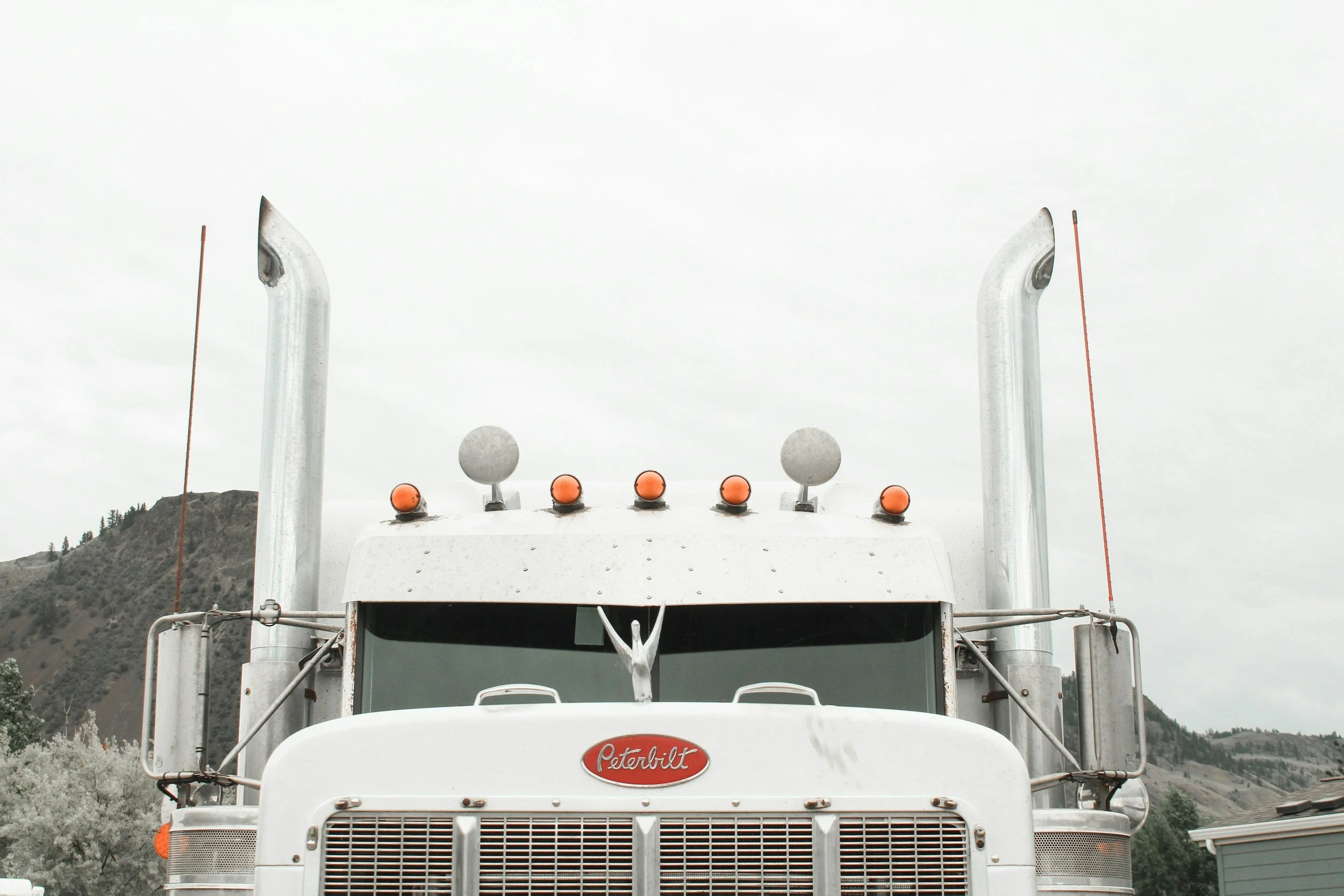 Front view of a Peterbilt semi-truck with a white cab, featuring multiple orange warning lights on top and two tall vertical exhaust pipes, set against a mountainous landscape with cloudy sky.