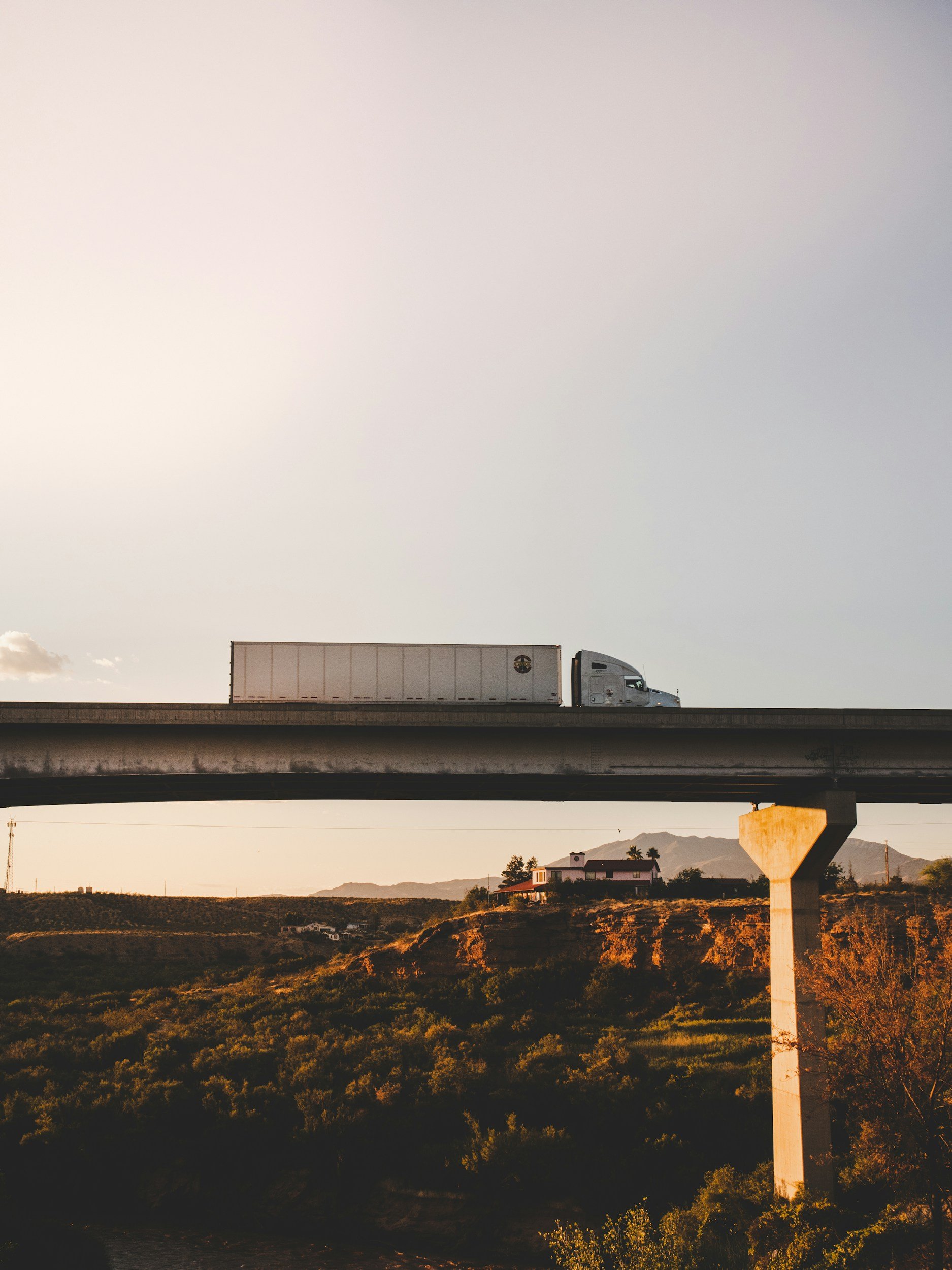 A white semi-truck driving on an overpass in a rural area during sunset, with mountains and houses in the background and trees in the foreground.