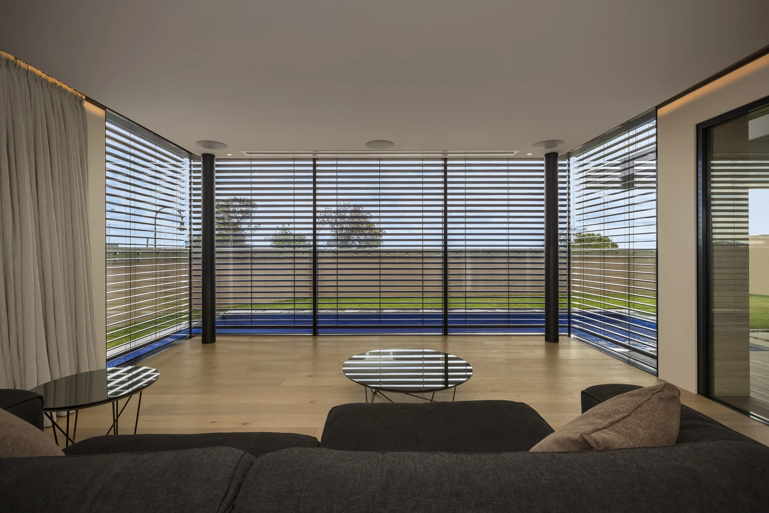 Living room with large windows with External Venetian Blinds, view of outdoor greenery, and minimal modern furniture including a round coffee table and sofa.