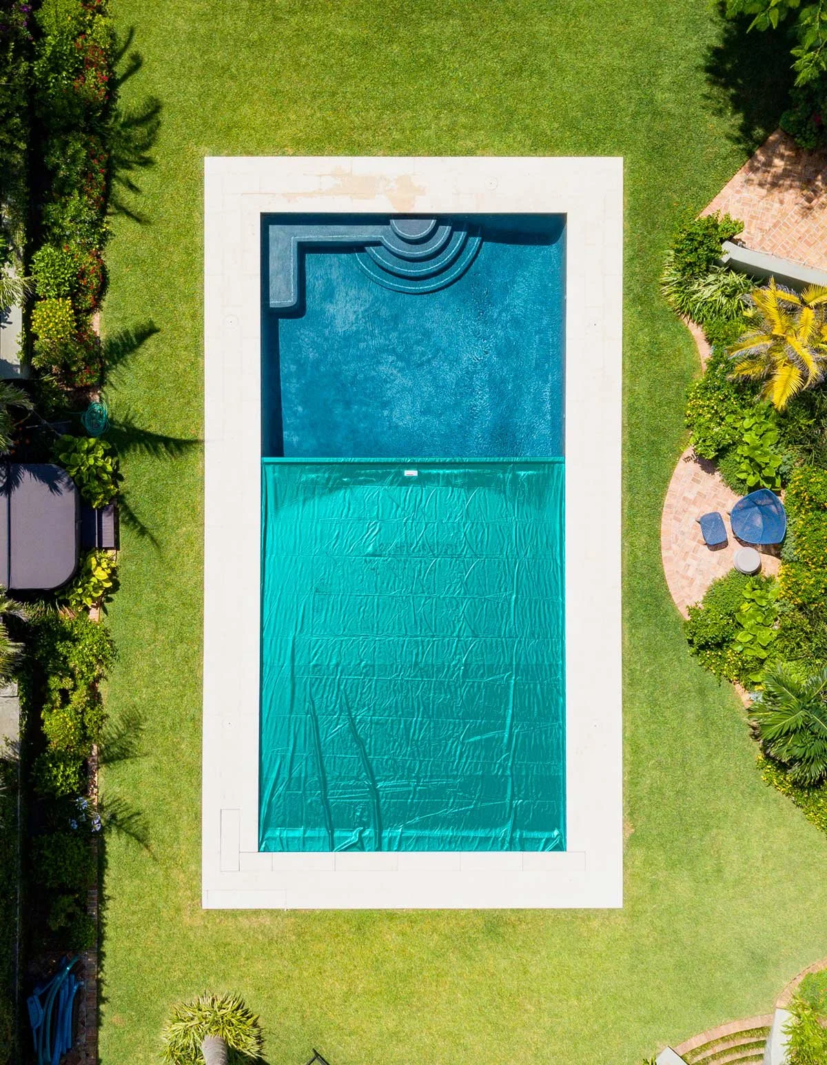 An aerial view of a rectangular swimming pool with a covered section at the bottom, surrounded by green lawn, trees, plants, and patio furniture.
